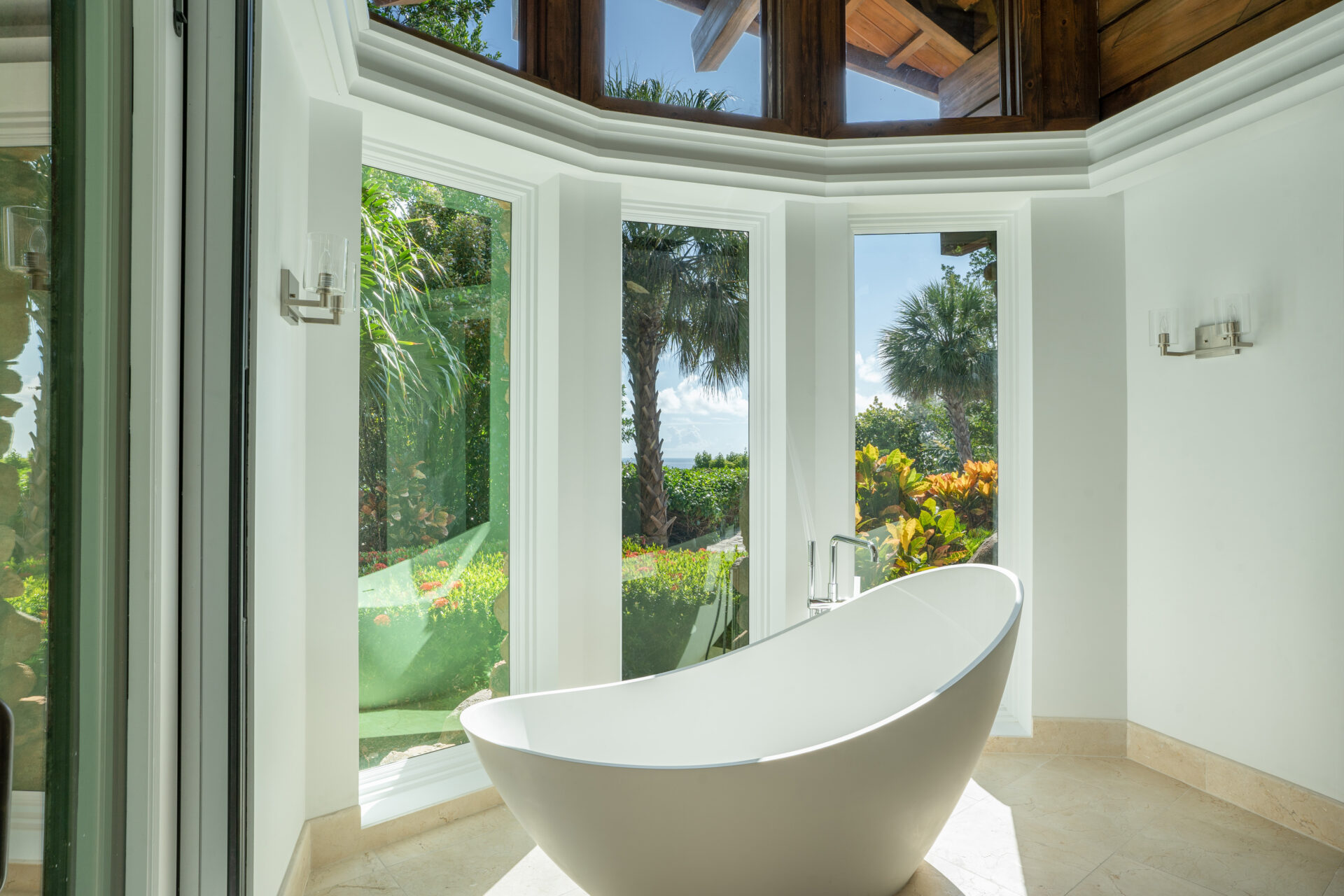 A modern, freestanding bathtub is placed in a bright bathroom with large, floor-to-ceiling windows. The windows offer a scenic view of lush green trees and clear blue sky, creating a serene and luxurious atmosphere reminiscent of a villa. The bathroom has white walls and a beige floor.