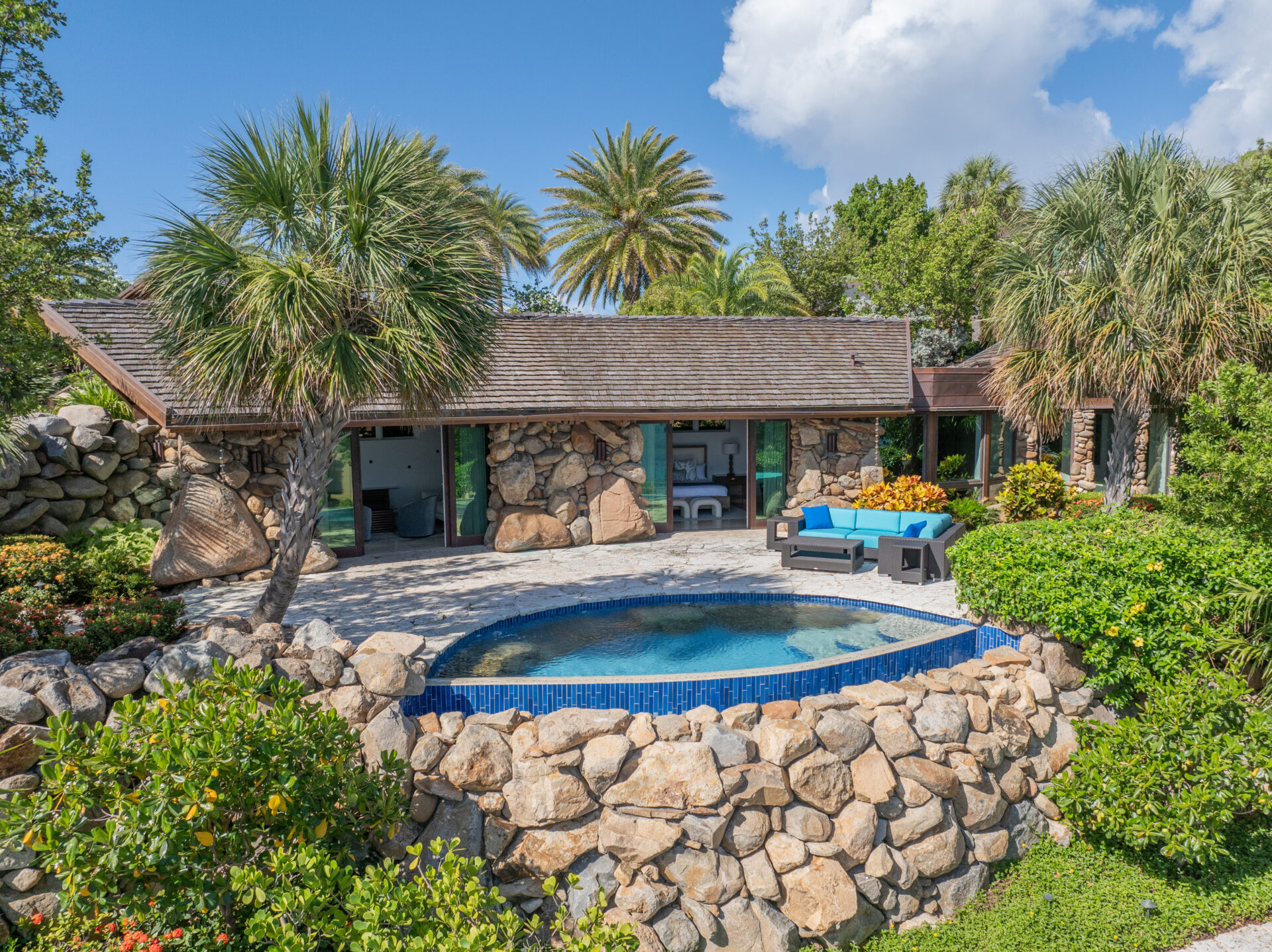 The luxurious Wings Villa features a stone house with a sloping wooden roof and large glass doors that open to a patio. The patio, adorned with a curved blue couch and table, overlooks a small, round swimming pool. Lush greenery and tall palm trees surround the property under a partly cloudy sky.