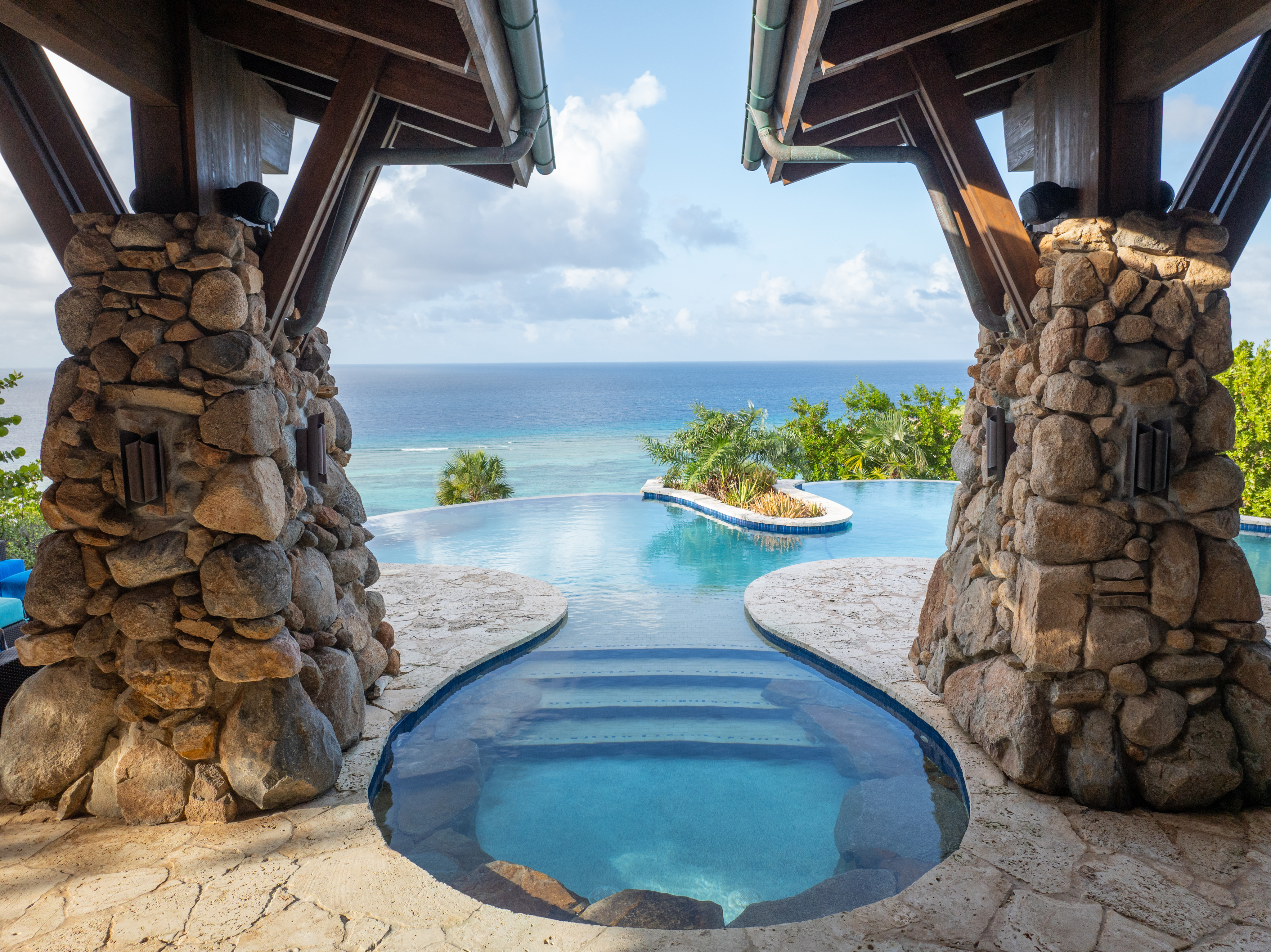 A scenic view of a luxurious infinity pool overlooking a tropical ocean. The Wings Villa pool area features rustic stone columns and a curved, organic design leading into the larger pool. The ocean and sky are visible in the background, framed by lush greenery.