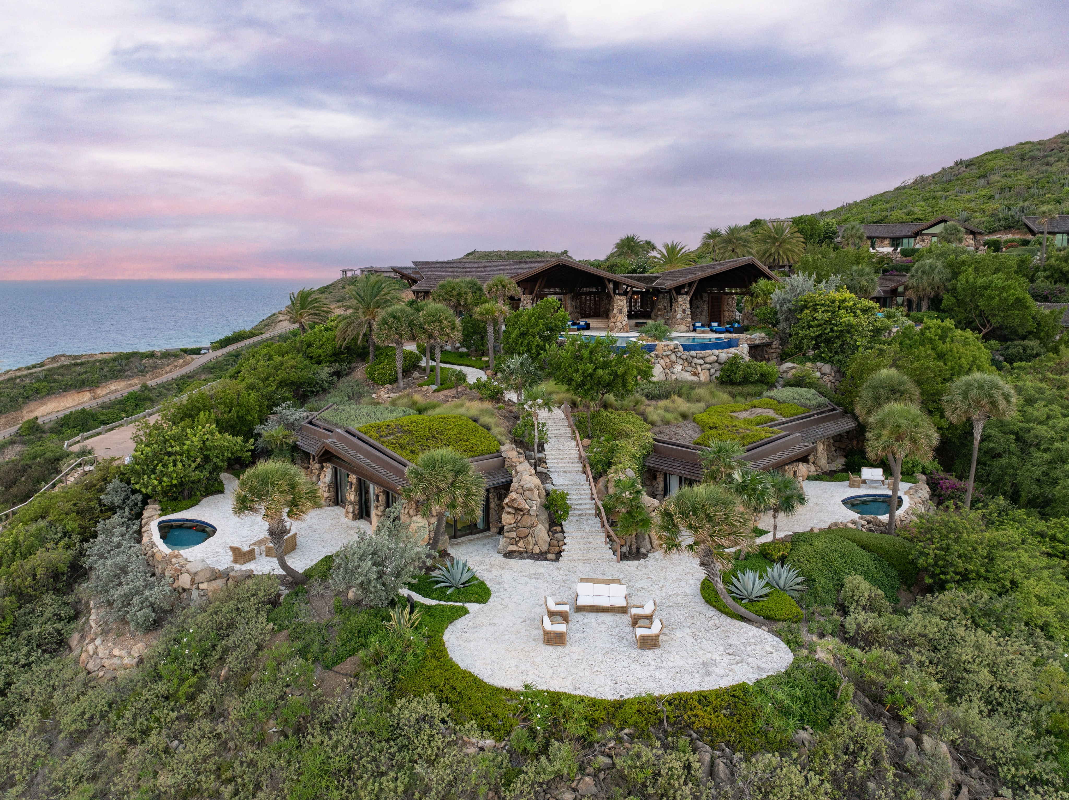 A luxury resort with multiple grass-roofed villas, including the stunning Wings Villa, set on a lush hillside overlooking the ocean. The property features winding paths, outdoor seating areas, palm trees, and small pools surrounded by greenery. The sky is partly cloudy with pink and purple hues.