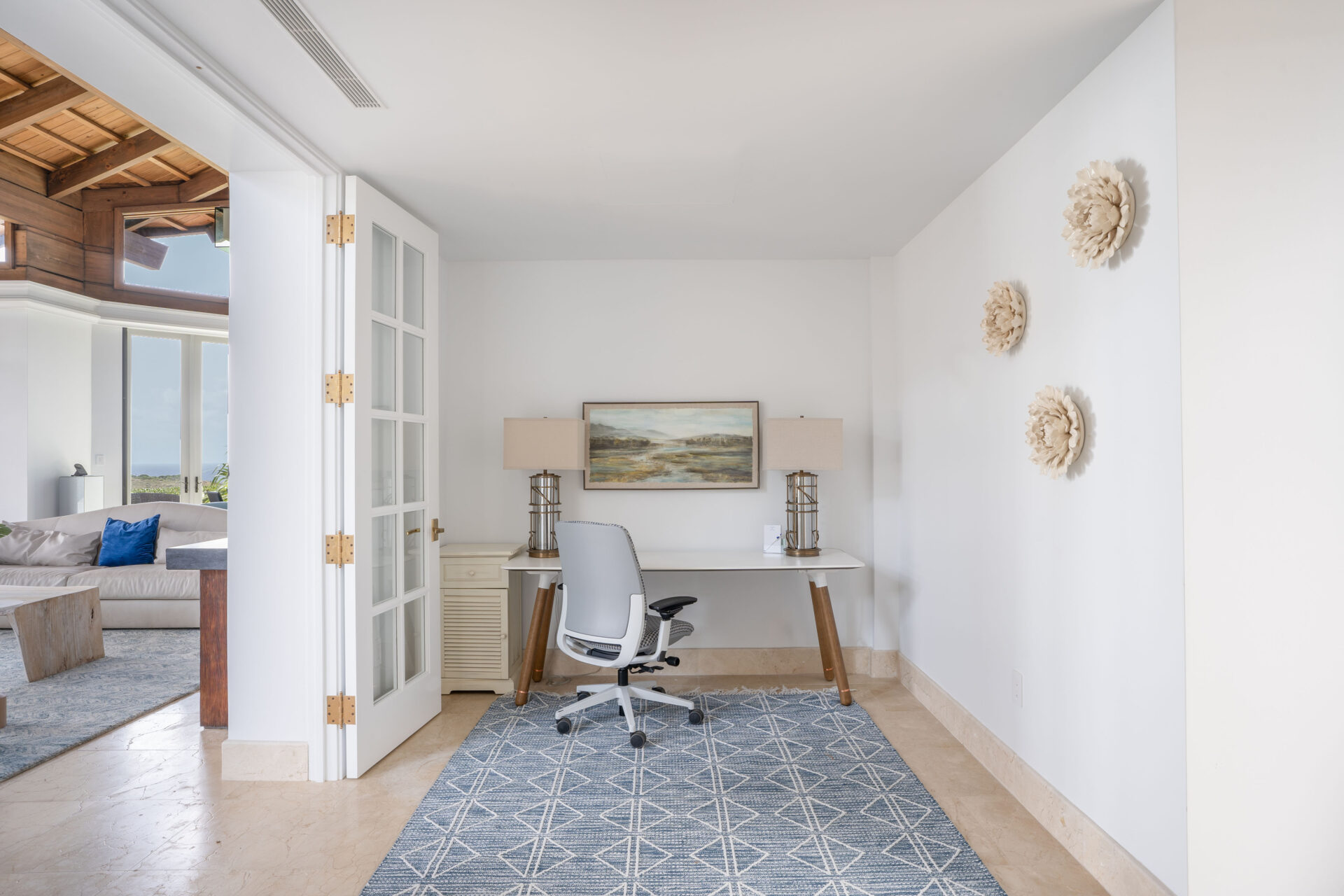 A minimalist home office in a villa, featuring a sleek white desk with a modern office chair, flanked by two beige lamps. A landscape painting hangs above the desk. The room has white walls, a geometric-patterned rug, and three decorative wall pieces. Wings of the adjoining living room are visible.