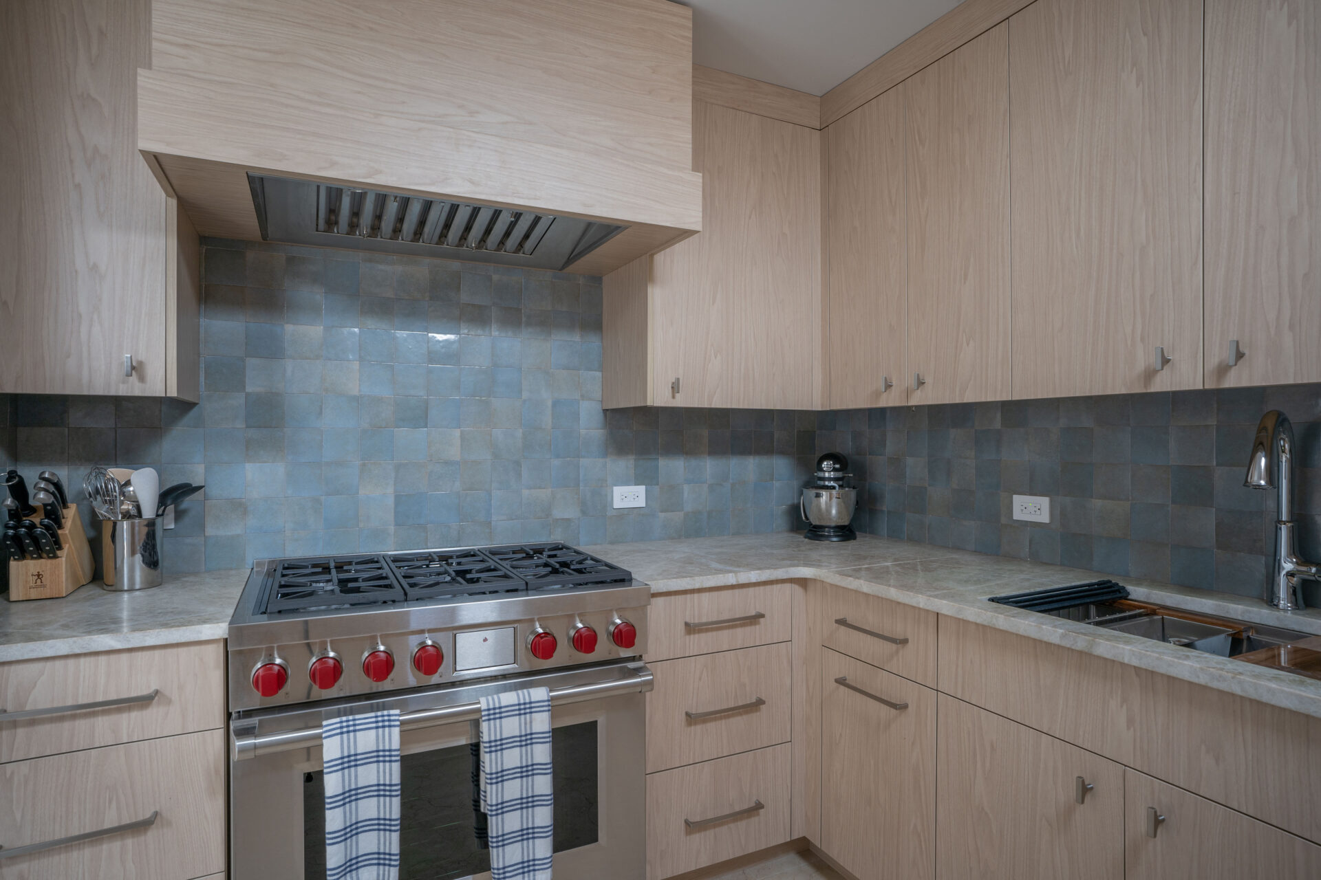 A modern kitchen in Wings Villa boasts light wood cabinets and a beige countertop. Below the vent hood, a stainless steel gas stove with red knobs stands out against a blue-tiled backsplash. A mixer, knife block, and sink are also visible, with two dish towels hanging on the oven handle.