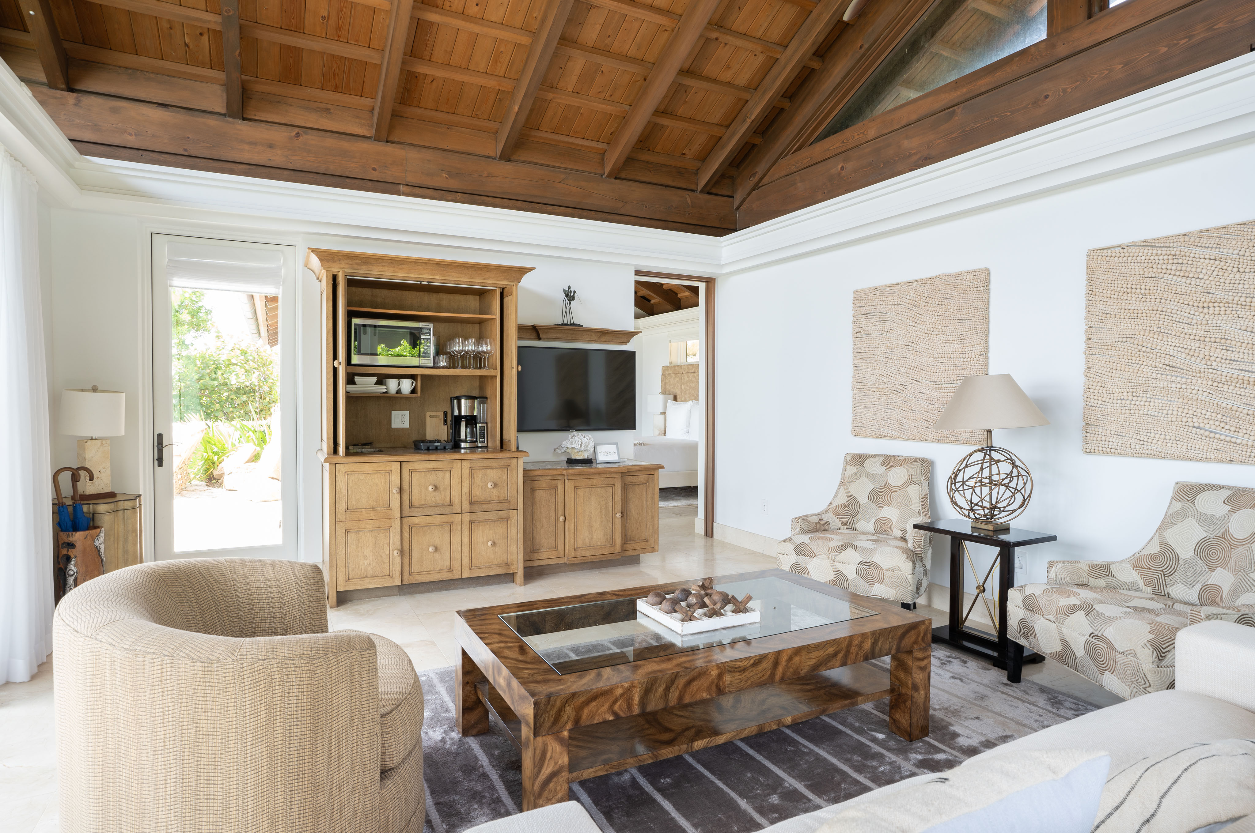 A cozy living room with a high wooden ceiling, beige and brown furniture, and a glass coffee table. Two patterned armchairs flank a side table with a decorative lamp. An entertainment center with a TV stands opposite, and a door leads to the bright outdoor area of the villa.