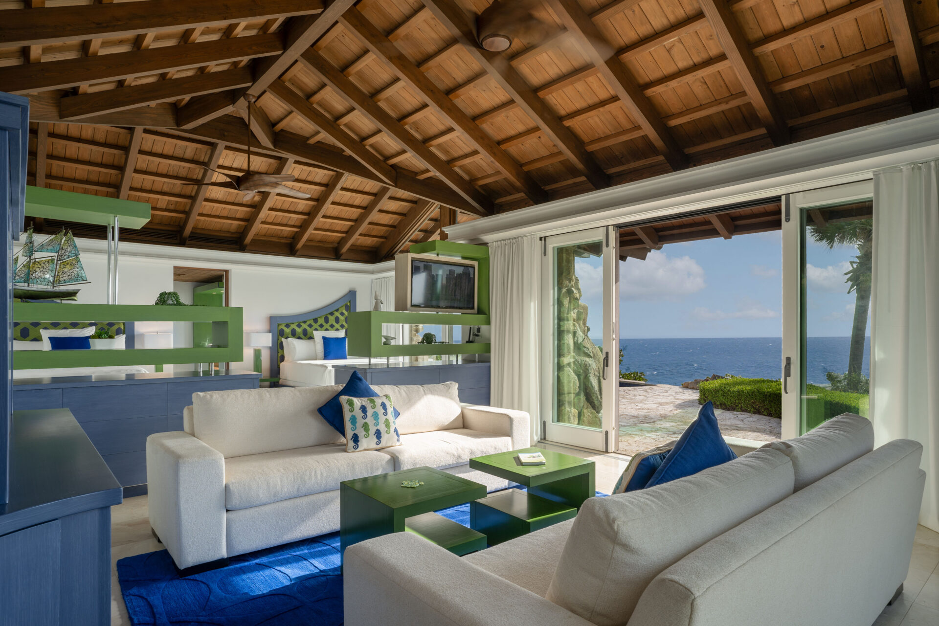 A spacious living room with wooden vaulted ceilings, containing beige sofas, a green and blue coffee table, and blue decorative accents. Large glass doors open to a stone patio with an ocean and clear sky view, allowing natural light to fill the villa's elegant wing.