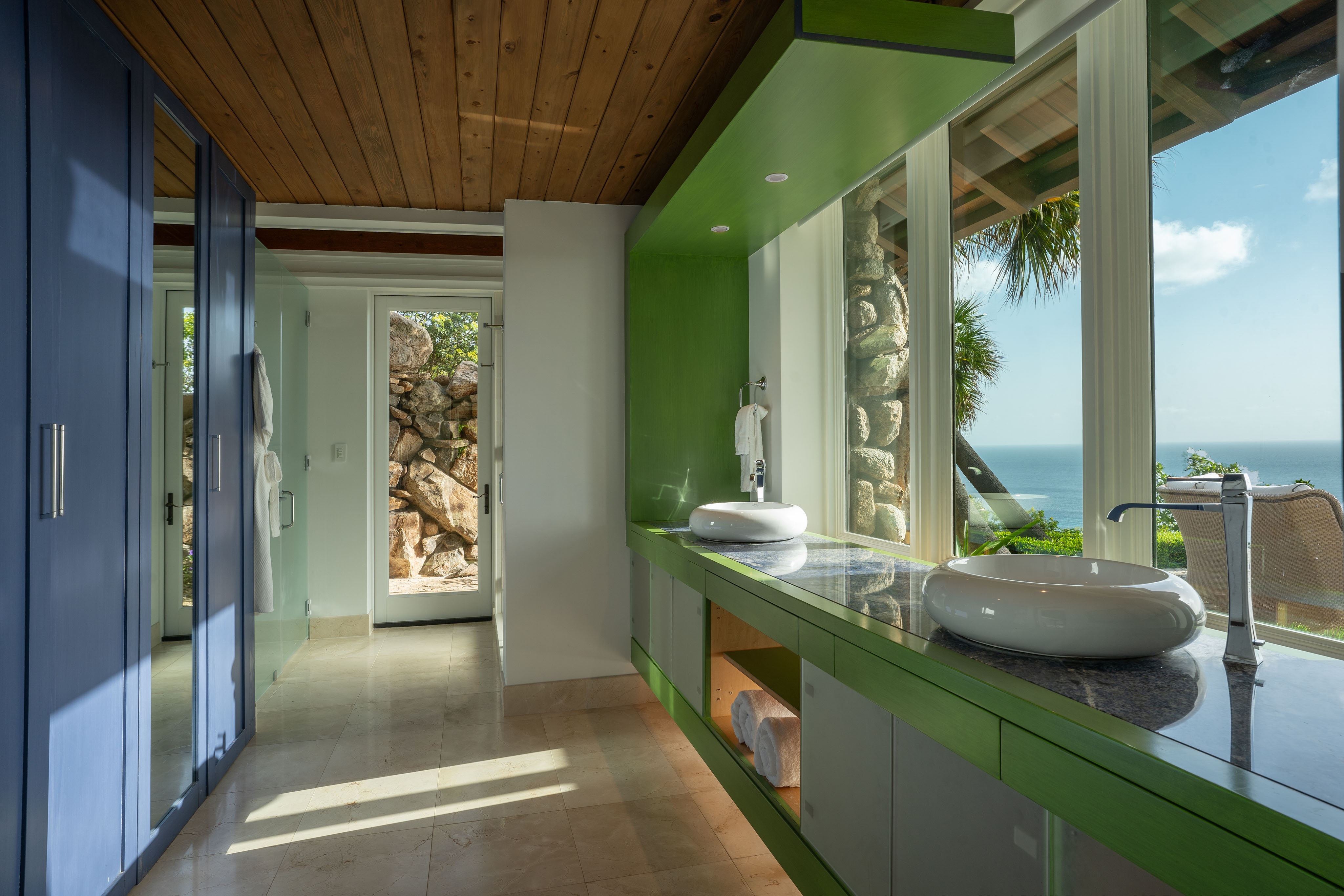 Spacious bathroom with wooden ceiling and large windows offering an ocean view in this stunning villa. Features a green countertop with two modern vessel sinks, blue cabinets, and a door leading outside. Sunlight fills the room, accentuating the serene and airy atmosphere like wings soaring in the sky.