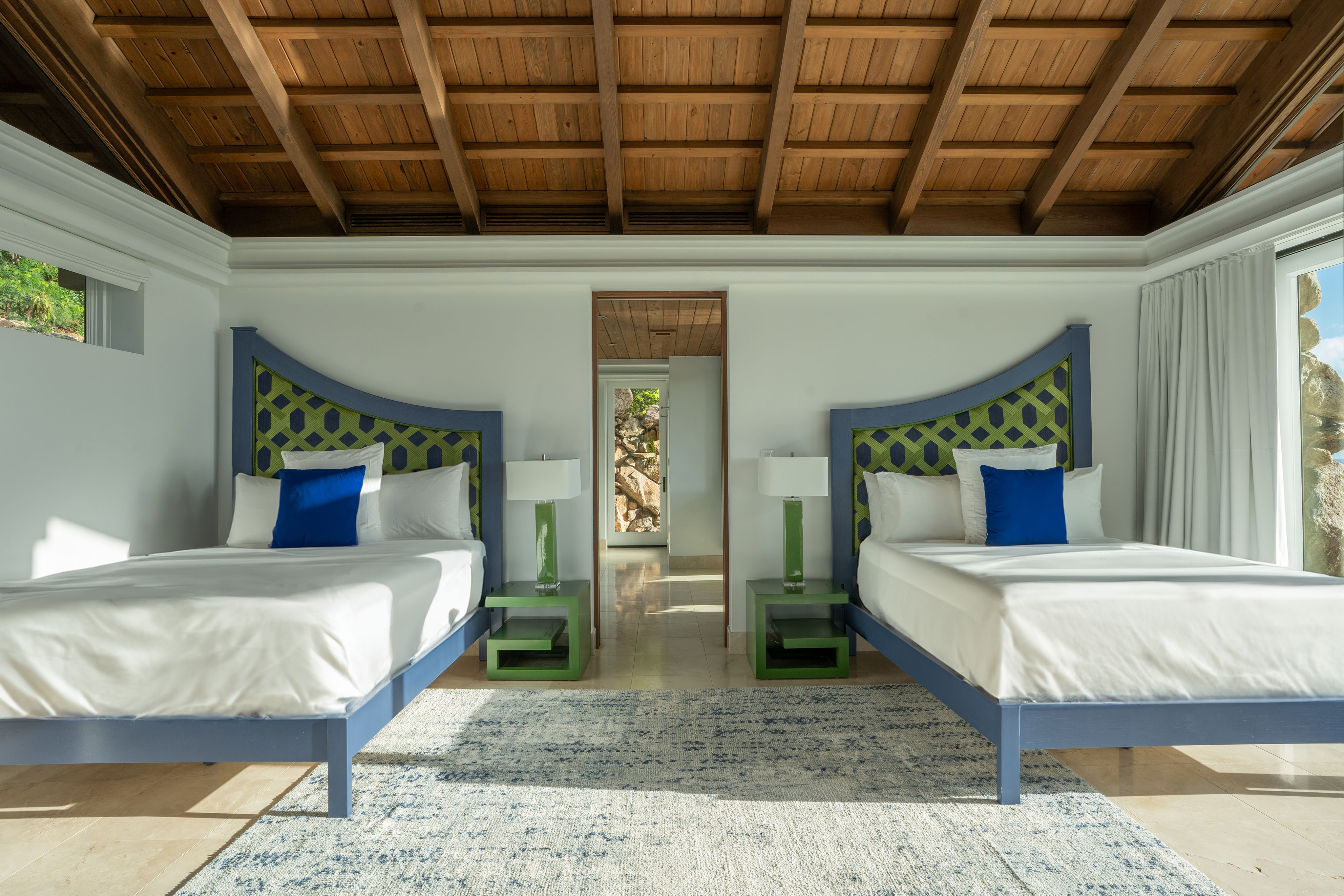 A well-lit bedroom in the villa with a high wooden ceiling. Two single beds with green and blue headboards, white linens, and blue accent pillows are positioned side by side. Between the beds are green nightstands with white lamps. A door in the back leads to another room, hinting at private wings beyond.