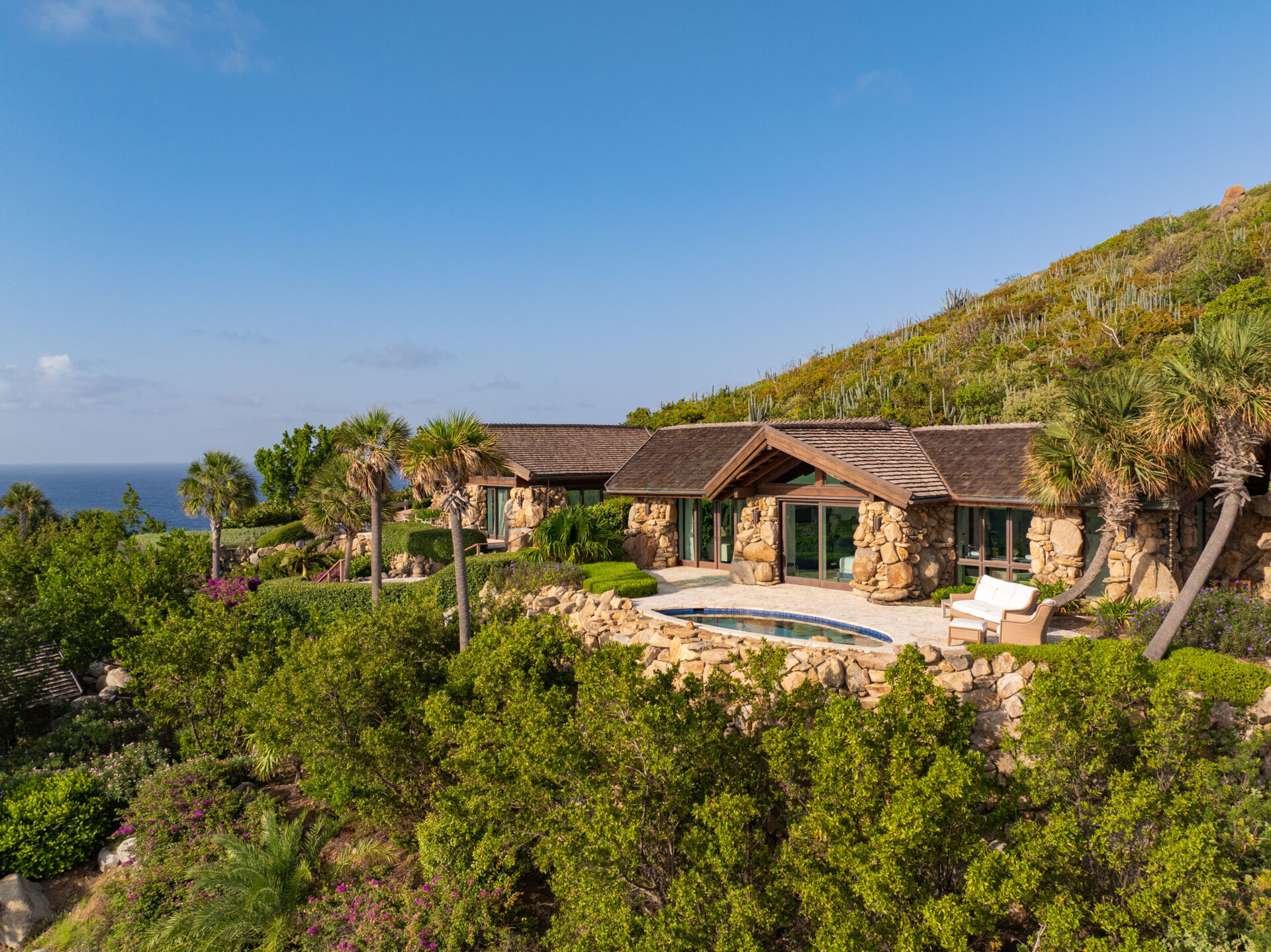 A stone villa overlooks the ocean from a lush hillside. The villa, topped with wooden roofs, features large windows and wings that extend to a round swimming pool on a terrace surrounded by greenery, palm trees, and blooming shrubs against a clear blue sky.