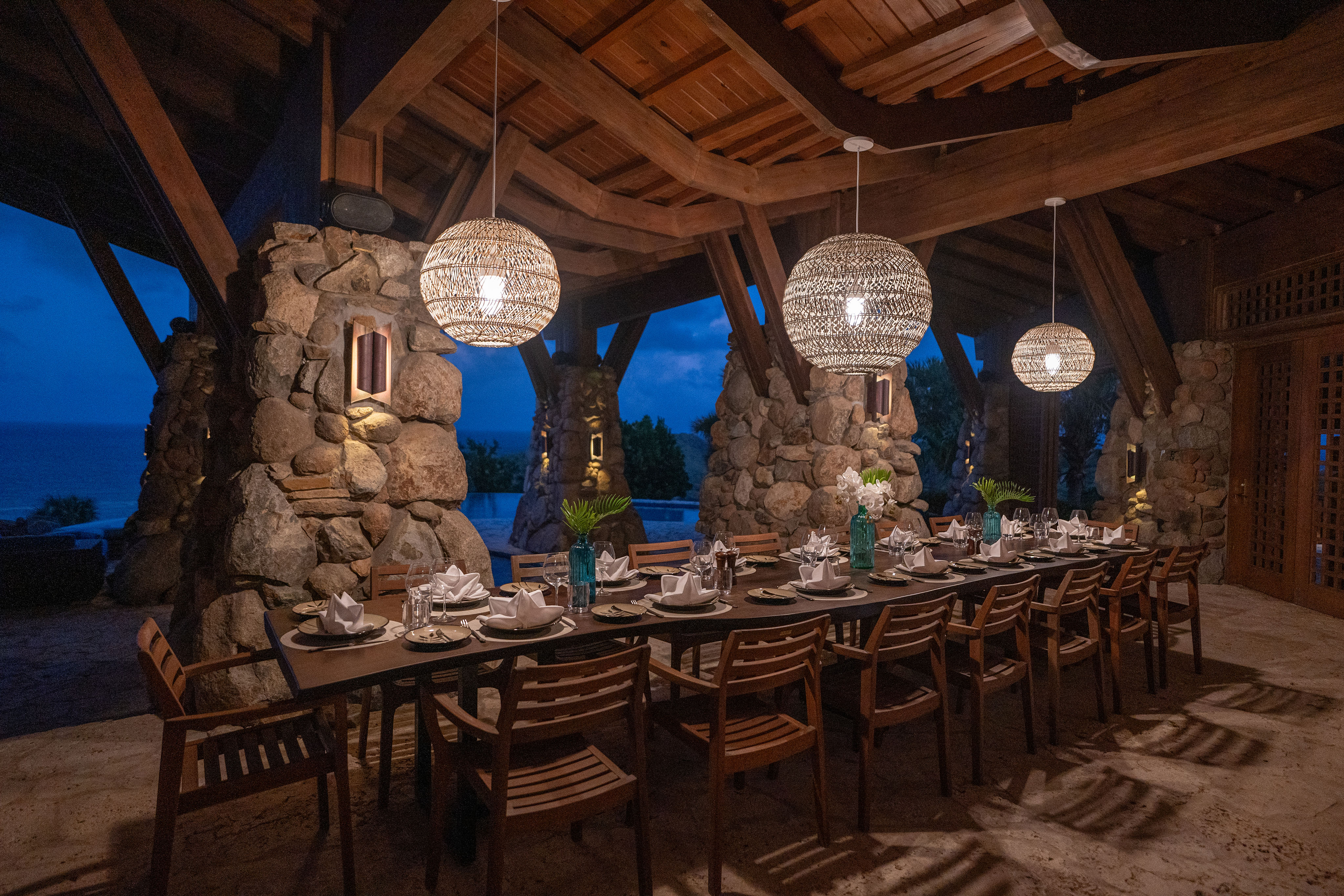 A long dining table set for dinner is positioned under a wooden pergola with stone pillars at Wings Villa. Three intricately designed hanging lanterns provide lighting. The setting is outdoors, with a view of the evening sky in the background, suggesting a serene, coastal location.