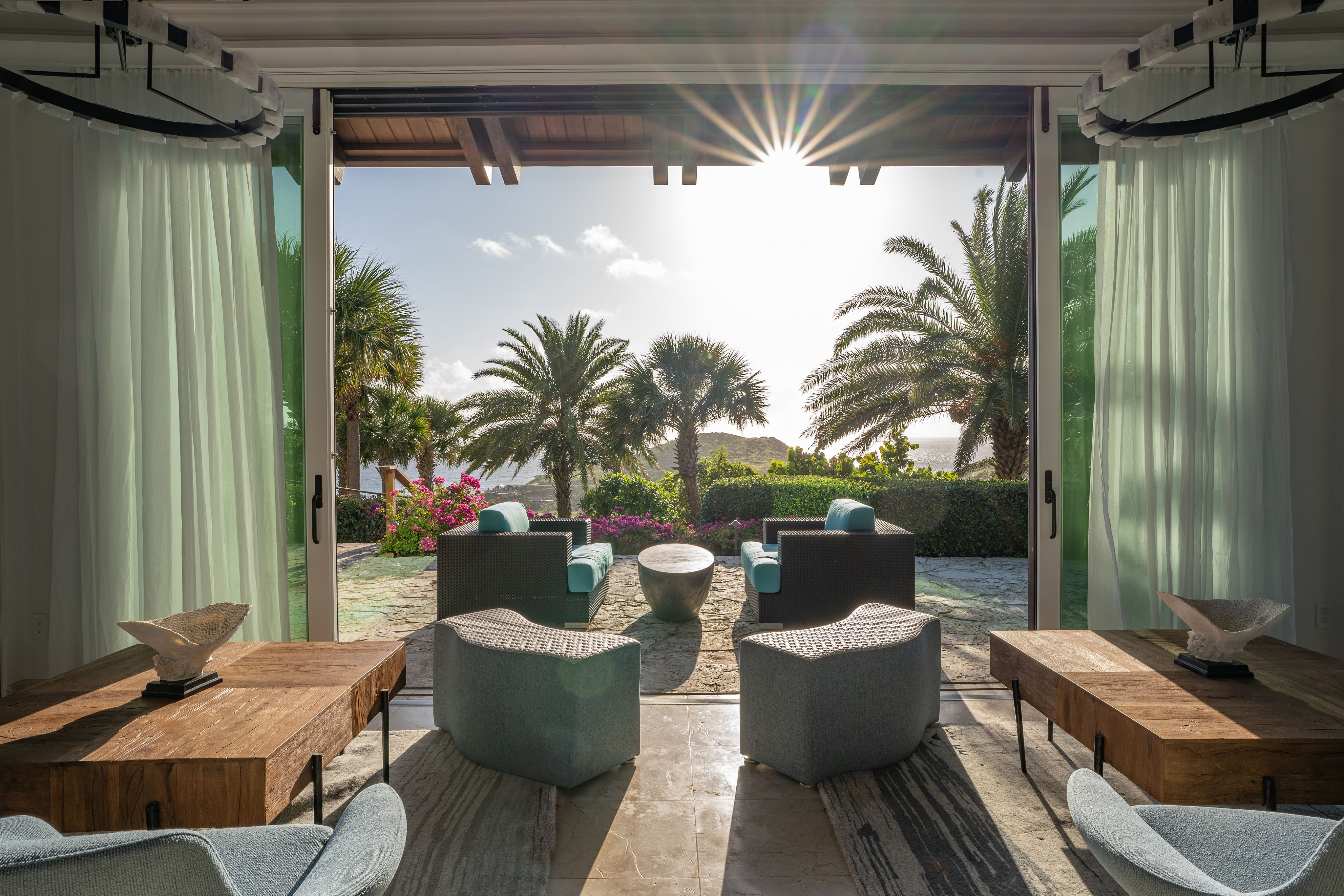 A sunlit, open-concept living area with modern furnishings extends to an outdoor patio. This villa's patio features a mix of cushioned seating and a central table, all framed by lush greenery and tropical palm trees, overlooking a serene ocean view, perfect for feeling like you're on wings.