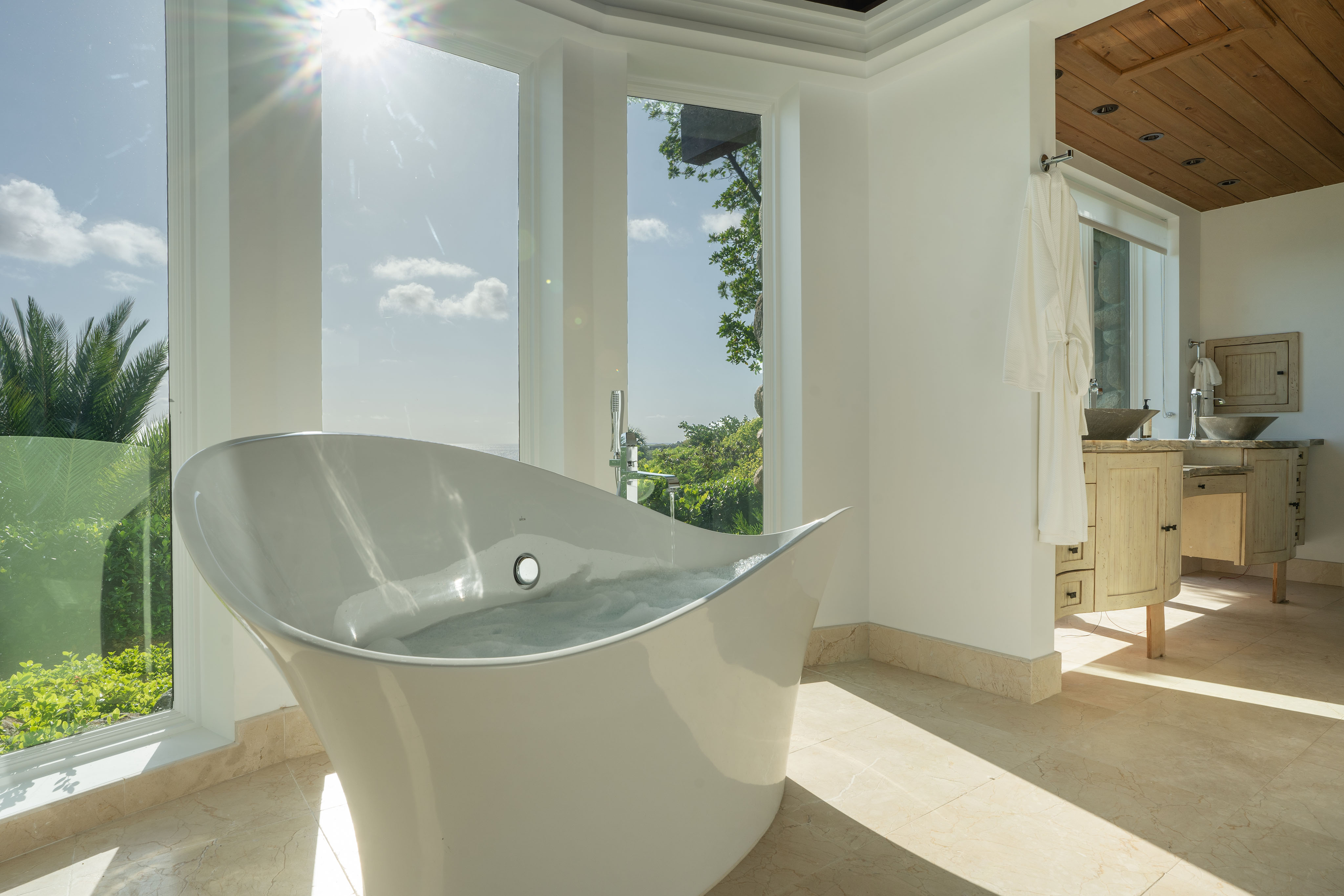 A spacious, sunlit bathroom in the villa features a modern, freestanding white bathtub positioned near large floor-to-ceiling windows. The bright space includes light-colored walls, a wooden vanity area with a sink, and lush greenery visible through the windows that seem to extend like wings.