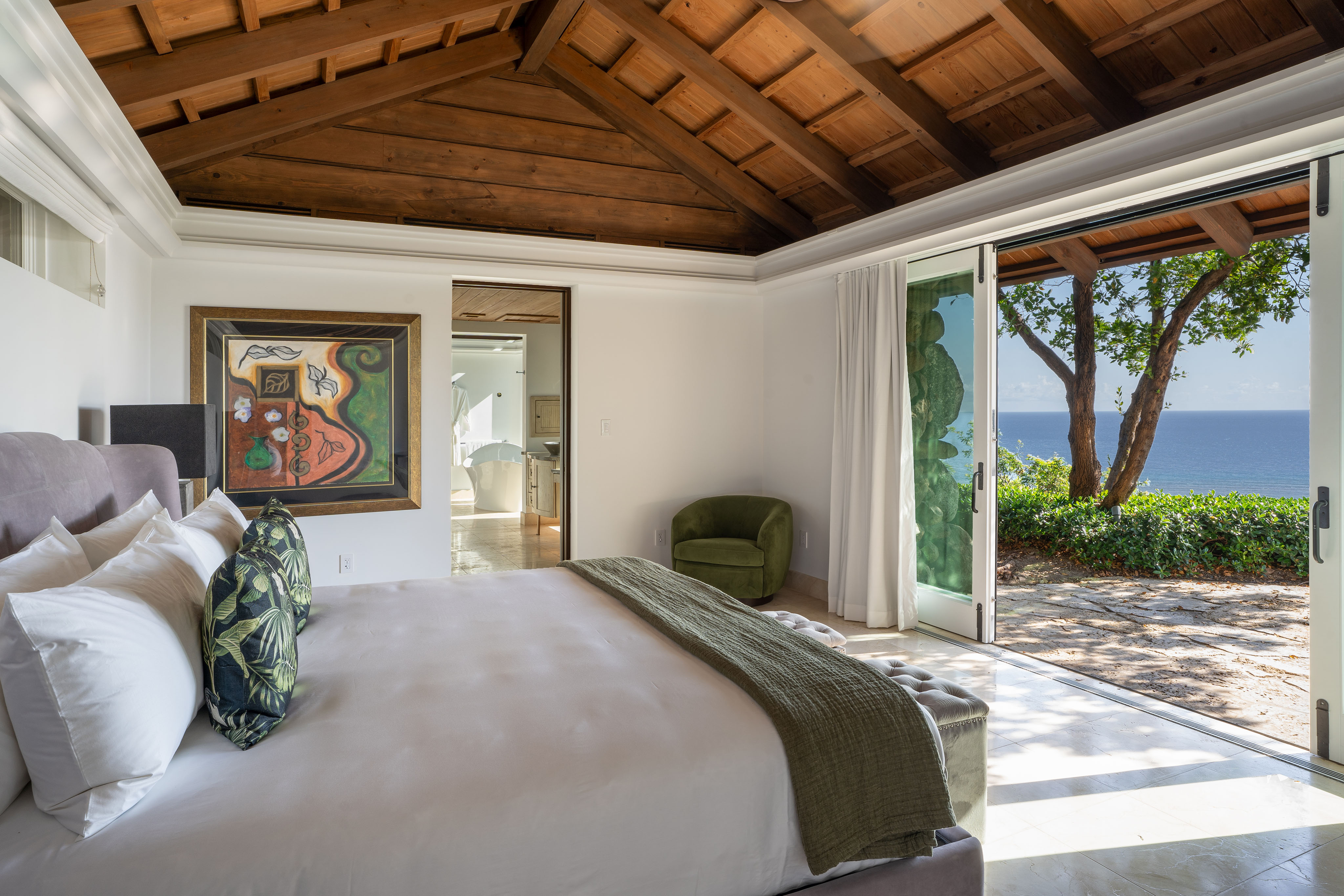 A luxurious villa bedroom with a vaulted wooden ceiling features a large bed adorned with white and green pillows and a green throw at the foot. The room has a green armchair, artwork on the wall, and glass doors opening to a patio with views of the ocean, offering an elegant retreat.