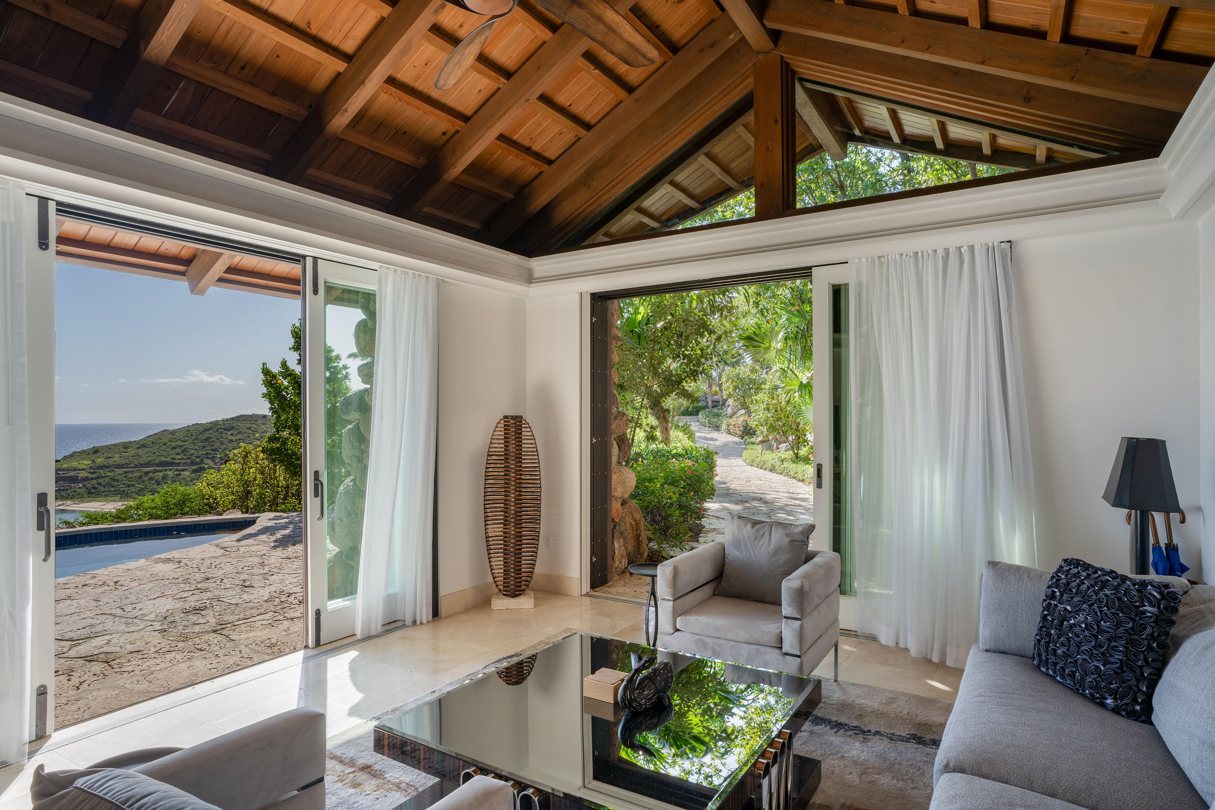 Luxurious living room in the villa, with large sliding glass doors opening to a scenic outdoor area. The room has high wooden ceilings, white walls, a beige sofa, a glass coffee table, and a wicker floor lamp. Outside, there's a view of lush greenery, a stone patio, and a pool that gives the space wings to nature.