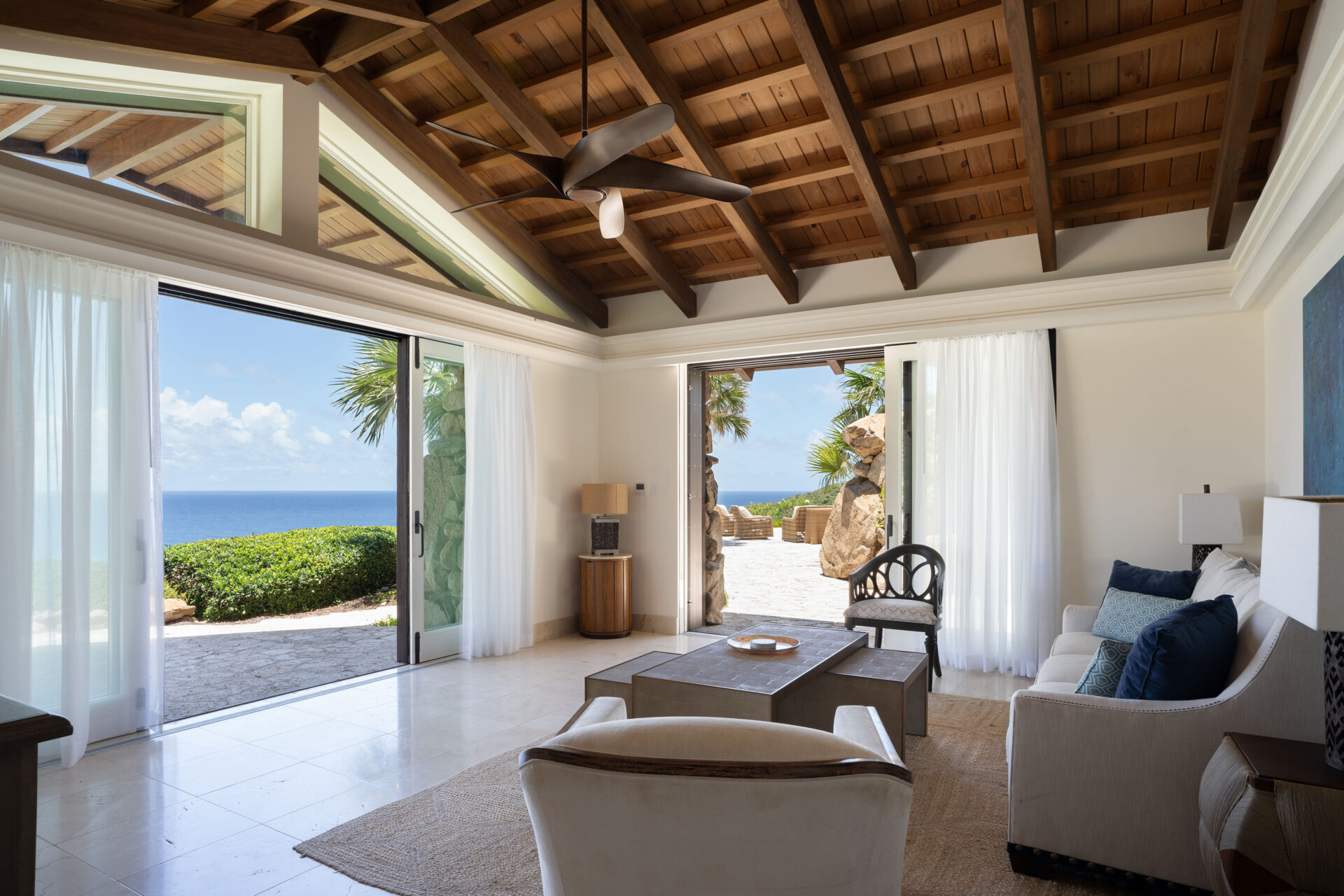 A spacious, airy living room in the villa features high, wood-beamed ceilings and large sliding glass doors opening to a patio with a view of the ocean. The room is furnished with a white sofa, chairs, a coffee table, and a side table. Light curtains frame the doors, letting natural light flood in.