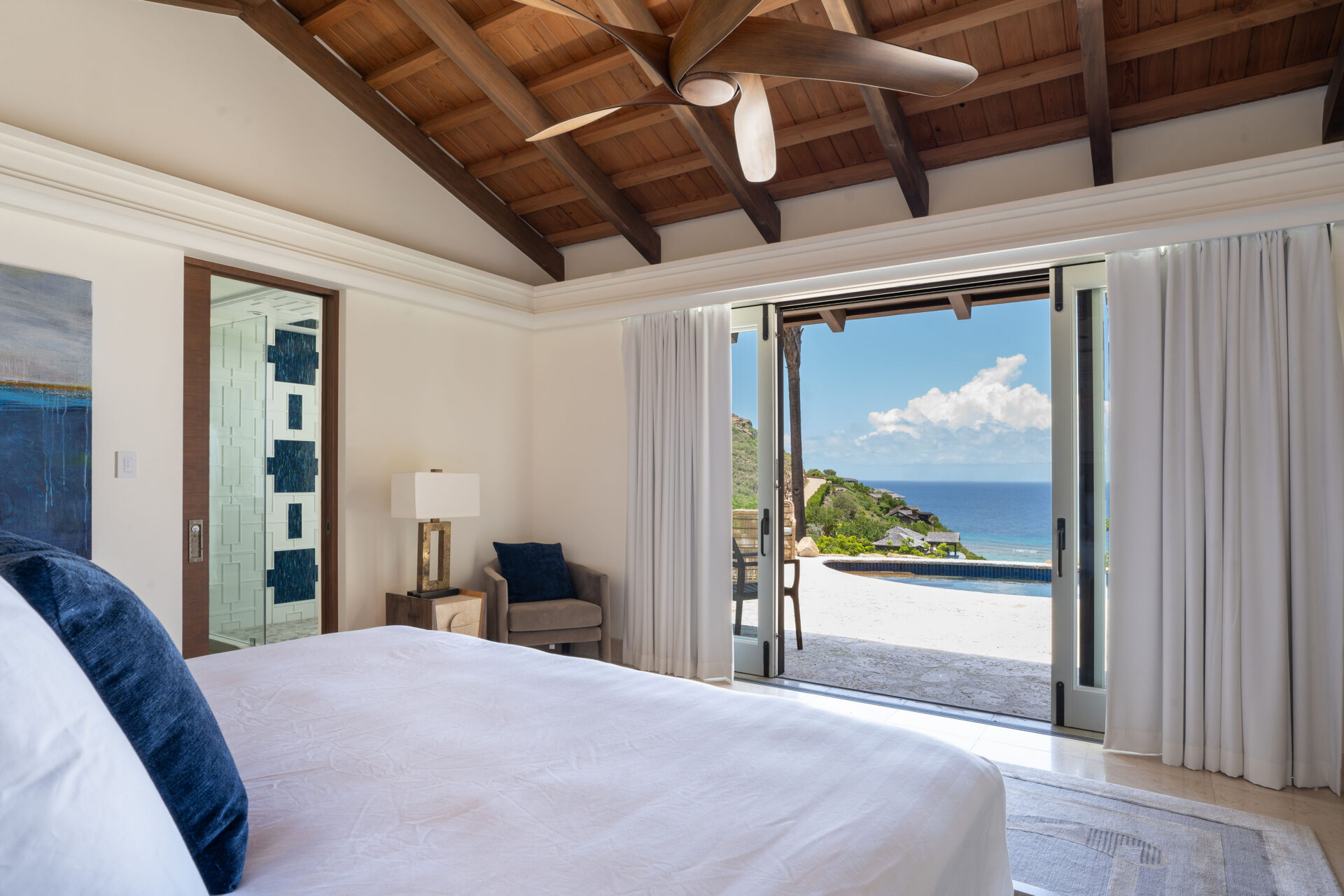 A serene bedroom with a high wooden ceiling and a ceiling fan features a large bed with white linens. Glass doors open to a stunning ocean view with lush green hills and a pool. Light floods the villa, highlighting its airy and relaxing ambiance, giving the room wings to embrace tranquility.