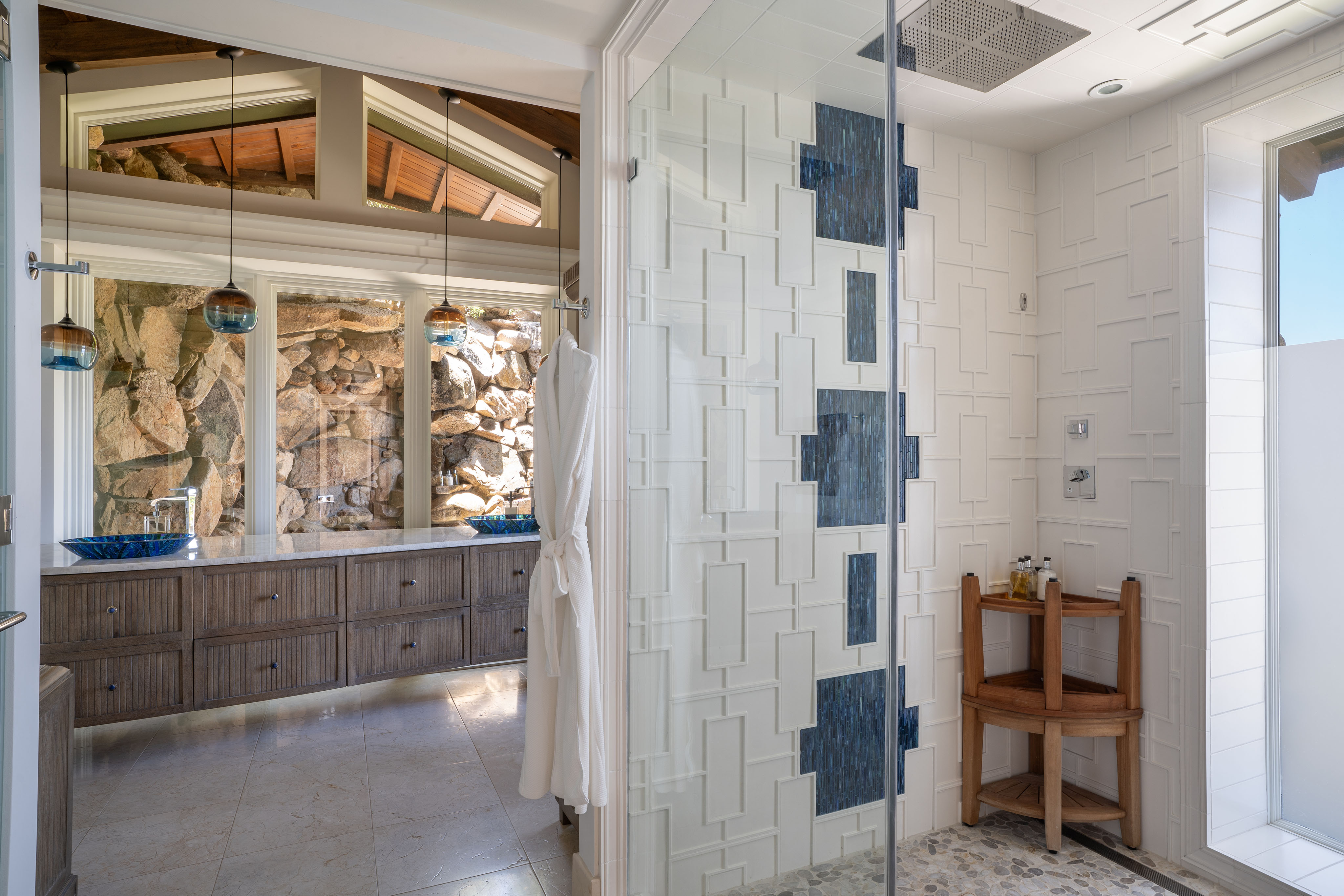 A modern bathroom in Wings Villa features a glass-enclosed shower with geometric white tiles and blue accents. Wooden elements, including cabinetry and a chair, add warmth. Two vessel sinks with blue bowl-shaped basins sit on a long counter, while natural light streams in from a large window.