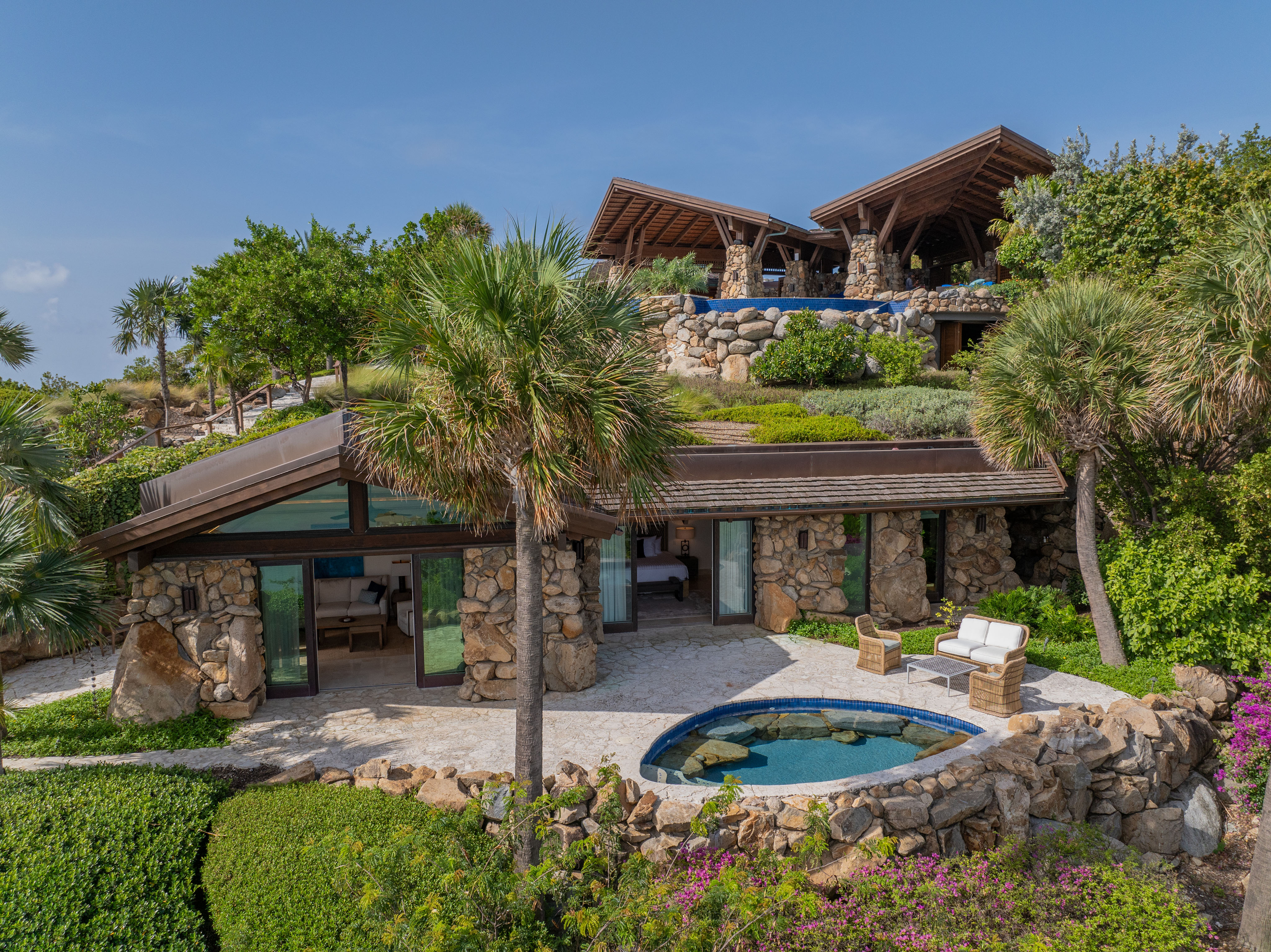 A luxurious stone villa with lush greenery, featuring a cozy patio with lounge chairs, a small circular pool, and large windows. The wing of the villa is nestled in a tropical setting under a clear blue sky, with another building visible on the hill above.