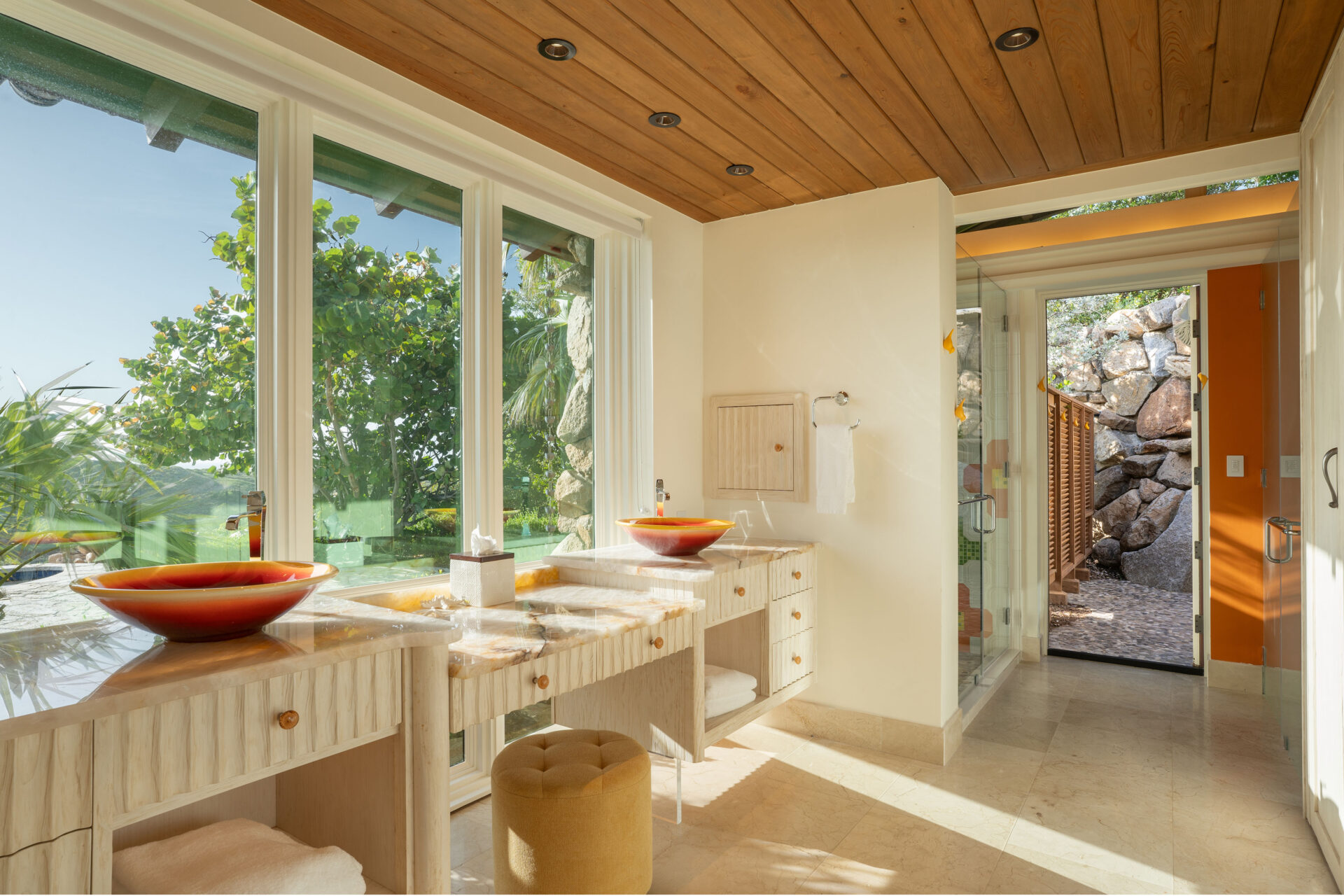 A spacious, well-lit bathroom in the villa, with large windows overlooking a green landscape. The room features two marble sinks with orange bowls, wooden cabinets, and a mirror. A wooden ceiling complements the natural light, and a glass shower door is visible in the background.