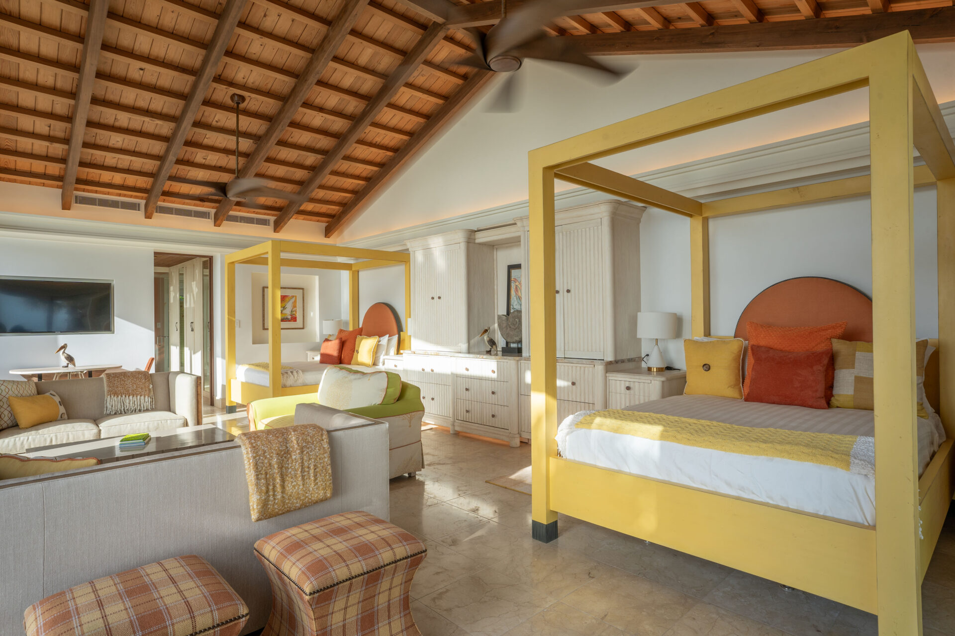 A spacious bedroom in the villa with a high, wooden beamed ceiling features two large, yellow canopy beds with colorful pillows and throws. The room includes a flat-screen TV, a seating area with sofas and chairs, and large windows allowing natural light to flood in.