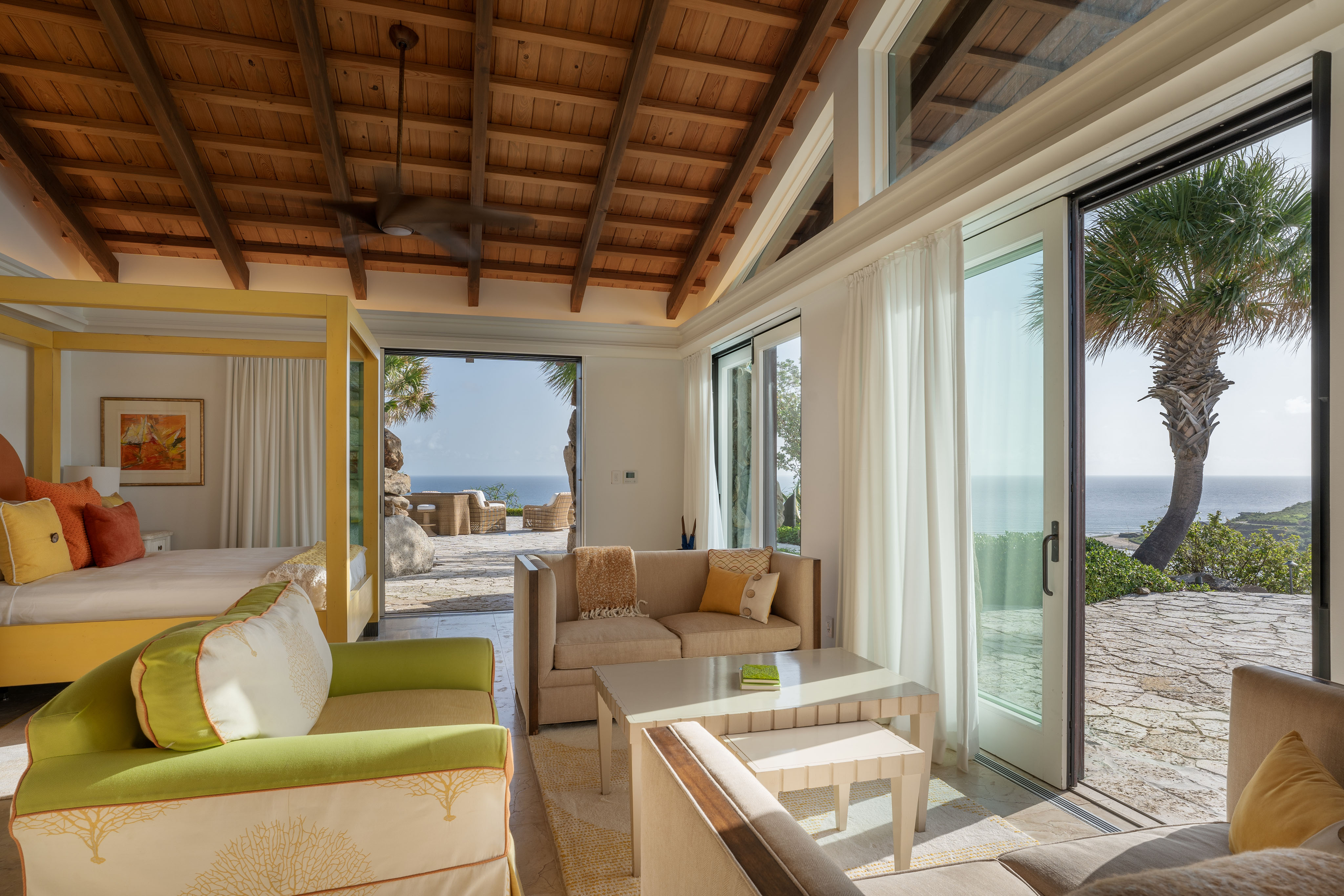 A spacious, sunlit living area in the villa features large sliding glass doors that open to a scenic patio overlooking the ocean. The room has a cozy sitting space with a beige sofa and green armchair, and a four-poster bed with yellow accents. Earthy tones and wooden ceilings enhance the ambiance.