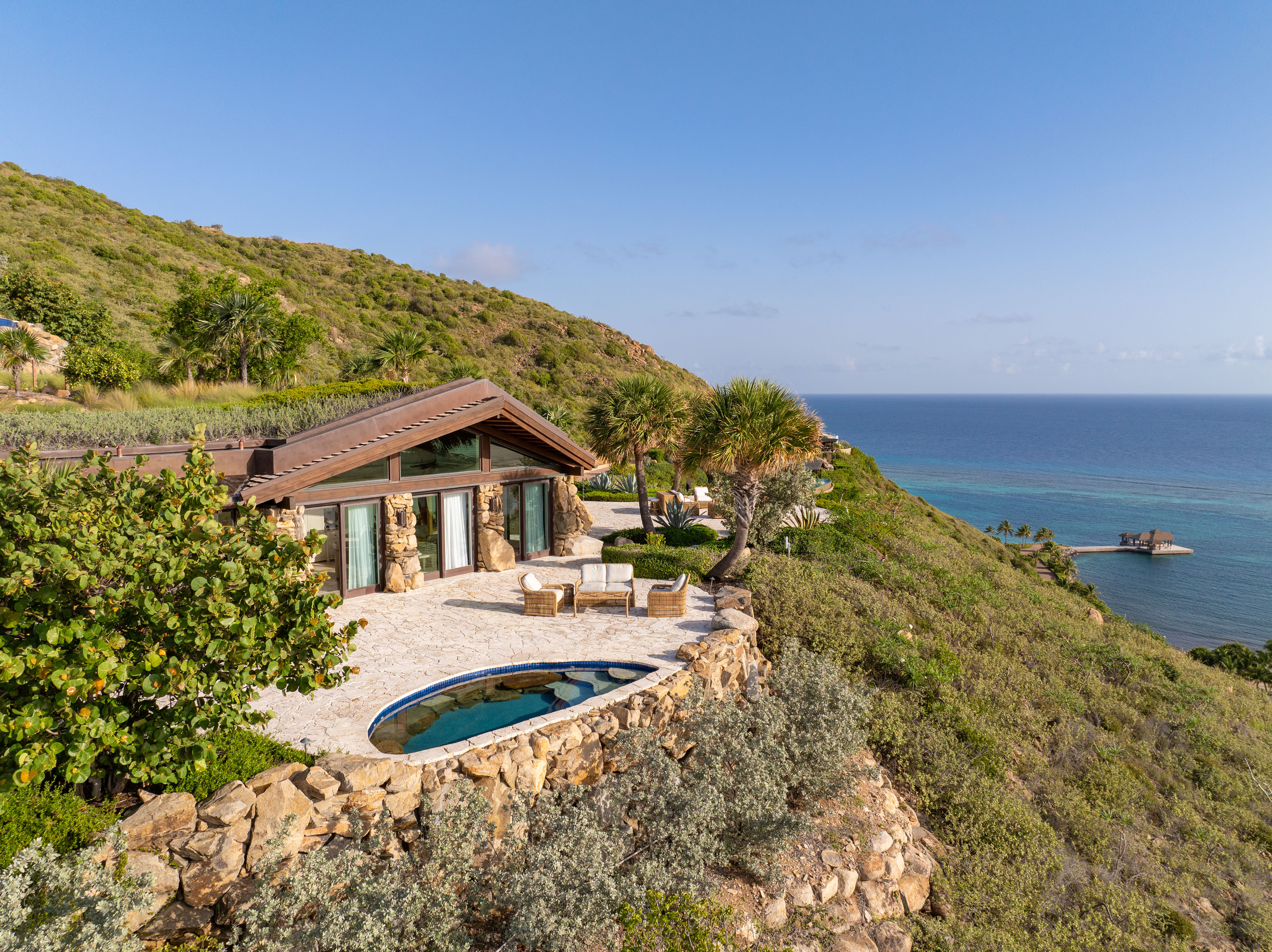 A luxurious hillside villa overlooking the ocean features a stone patio with outdoor seating and a small, round pool. The surrounding greenery and clear blue sky enhance the tranquil atmosphere, while an observation deck, spread like wings over the water in the distance, invites you to take in the breathtaking views.