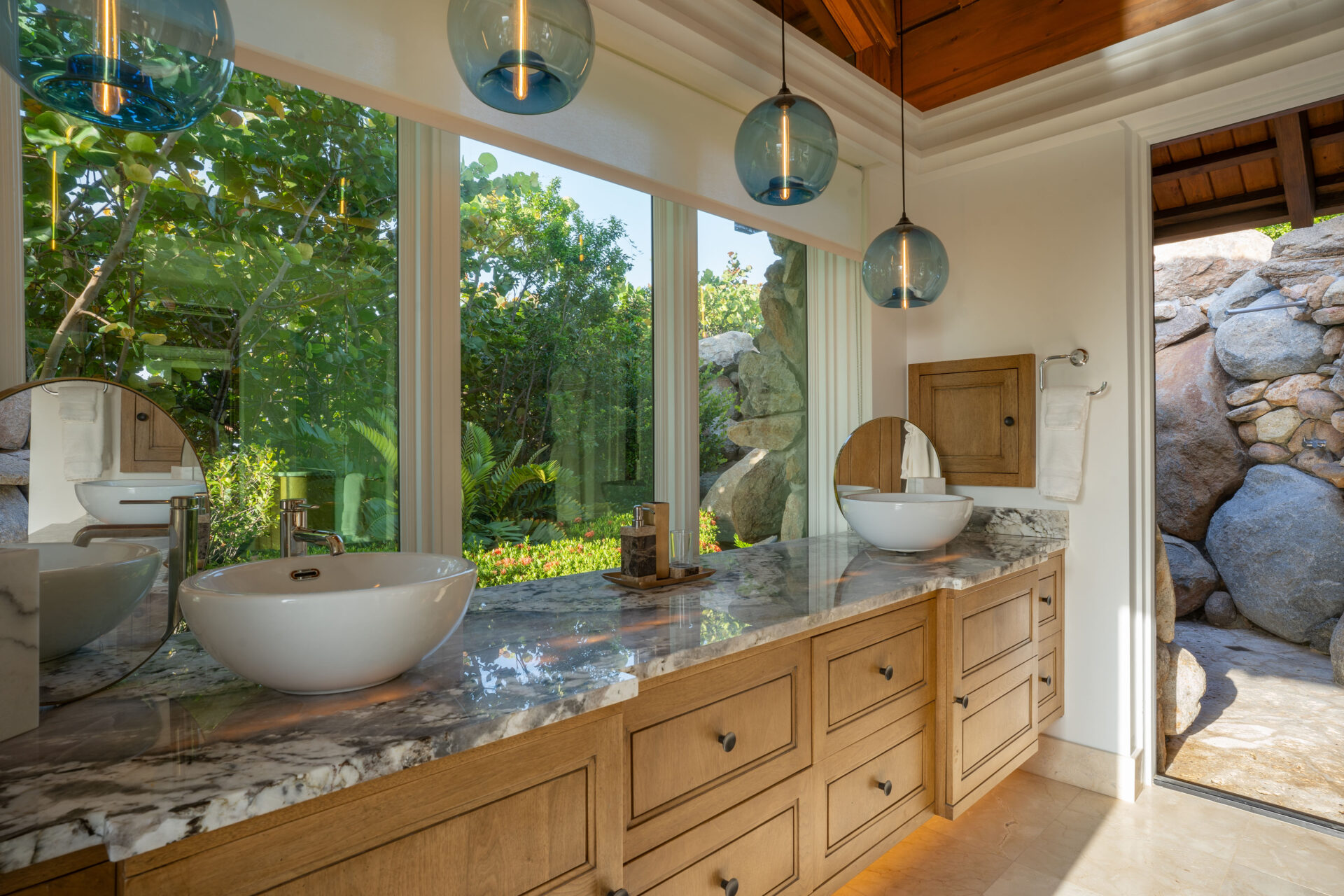 A luxurious bathroom in the villa boasts a large marble countertop, dual white vessel sinks, wooden cabinets, and round mirrors. Three blue glass pendant lights hang from the wooden ceiling. The space is bright with floor-to-ceiling windows offering a view of lush greenery outside, giving it wings to soar.