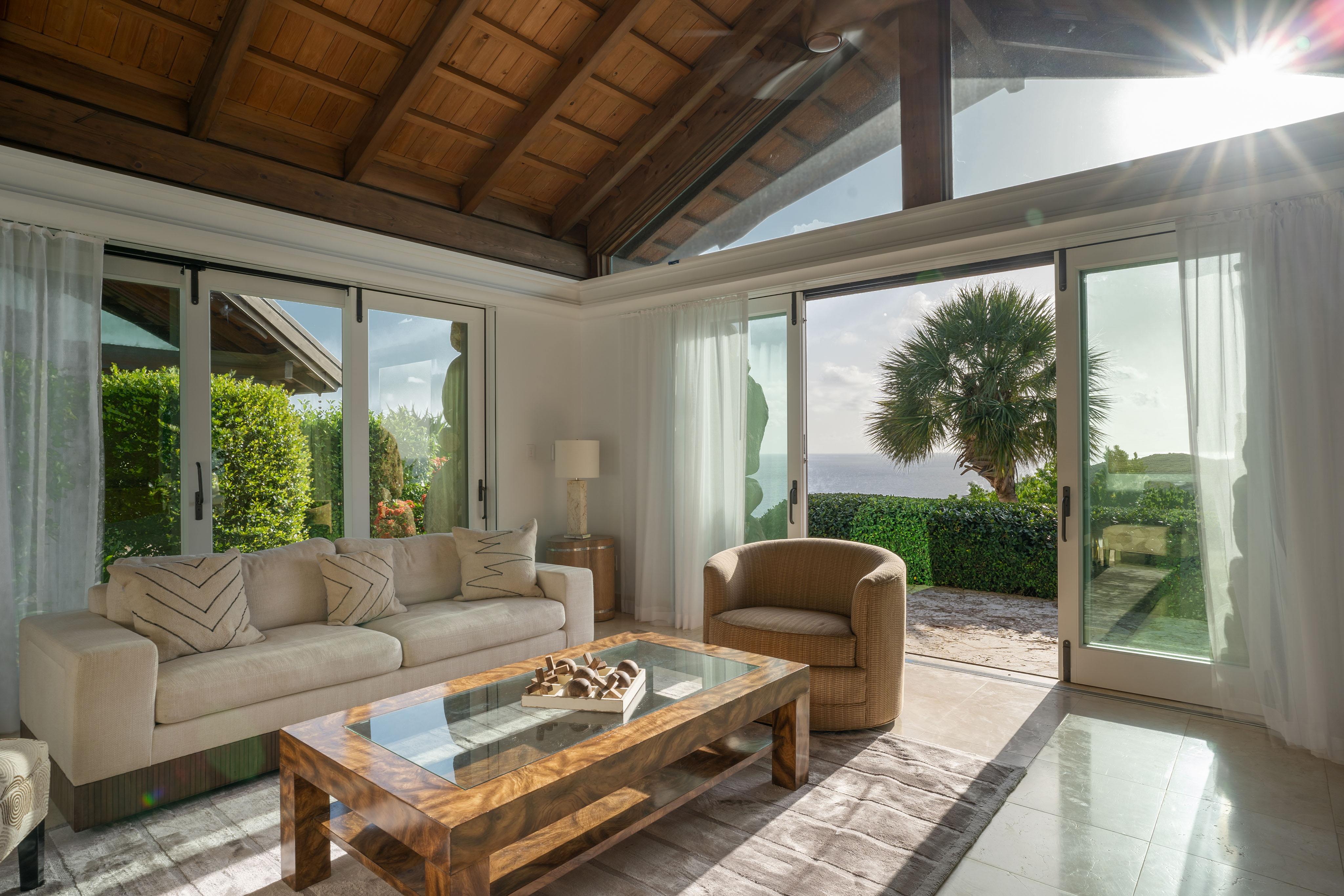 Sunlit living room with large windows and glass doors opening to an outdoor patio, showcasing a scenic view of the ocean and a palm tree. This villa features a beige sofa, a cushioned armchair, and a wooden coffee table on a tan tiled floor.