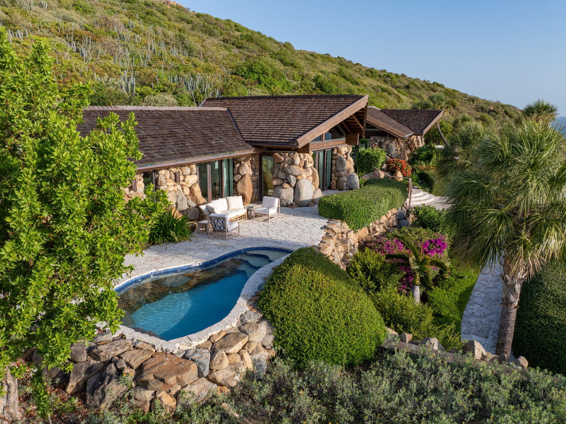 A picturesque villa with a stone exterior, nestled on a hillside amidst lush greenery. The house features a small, kidney-shaped pool surrounded by rocks and manicured bushes. There are outdoor seating areas with chairs and a coffee table, and wings that offer stunning views of the sea in the distance.