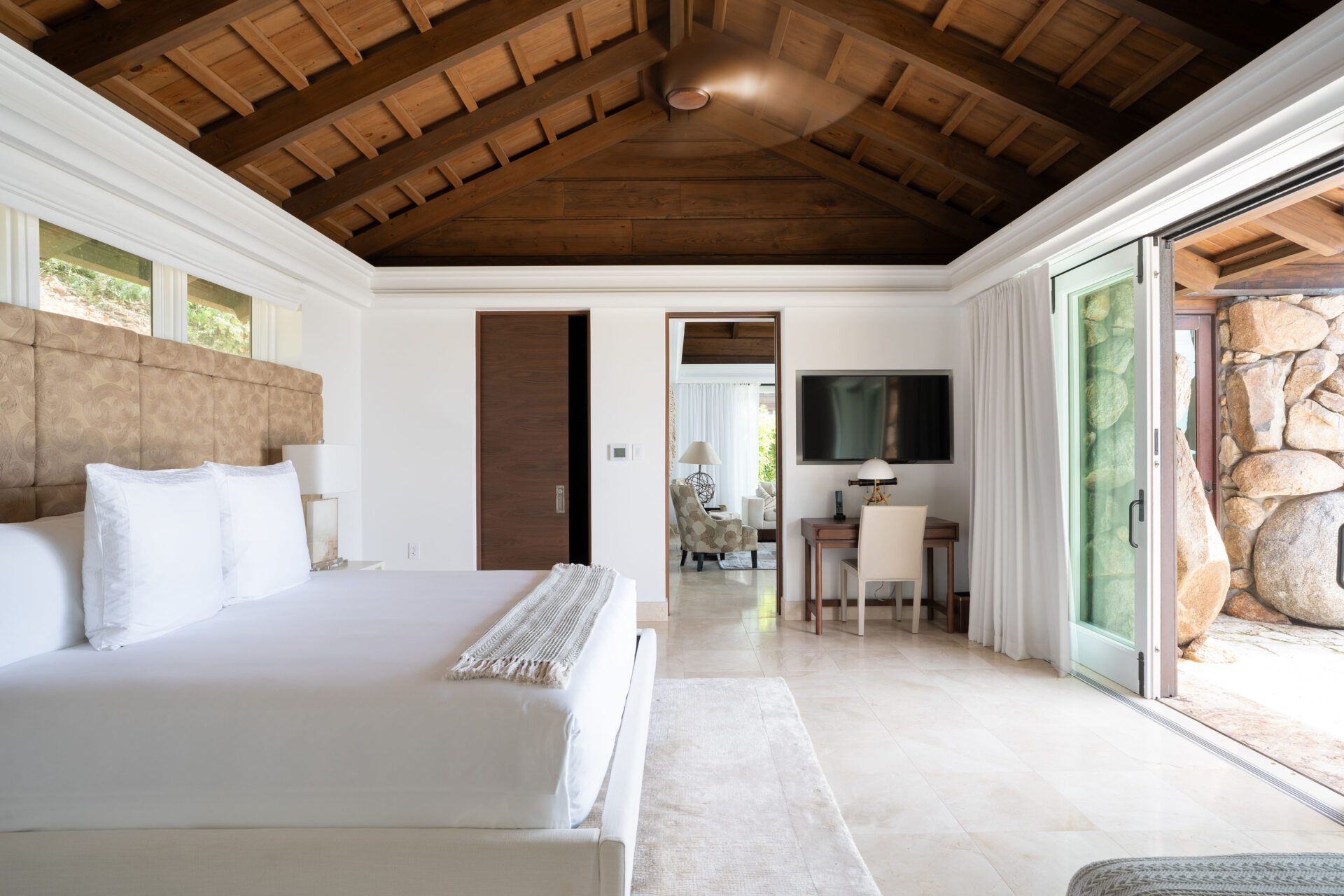 A spacious, modern bedroom in the villa features a high wooden ceiling, a large bed with white linens, a wall-mounted TV, a desk and chair, and sliding glass doors leading to an outdoor area. The room is bright and airy with natural light streaming in like wings of sunshine.