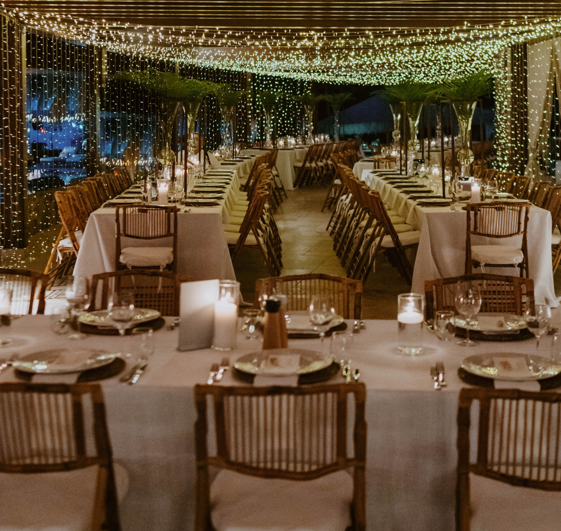 A beautifully decorated event space by BVI Weddings & Events with long tables set for a formal dinner. Each table features stylish tableware, glassware, and candles. Overhead, twinkling fairy lights drape down, casting a warm, inviting glow on the elegant rattan chairs below.