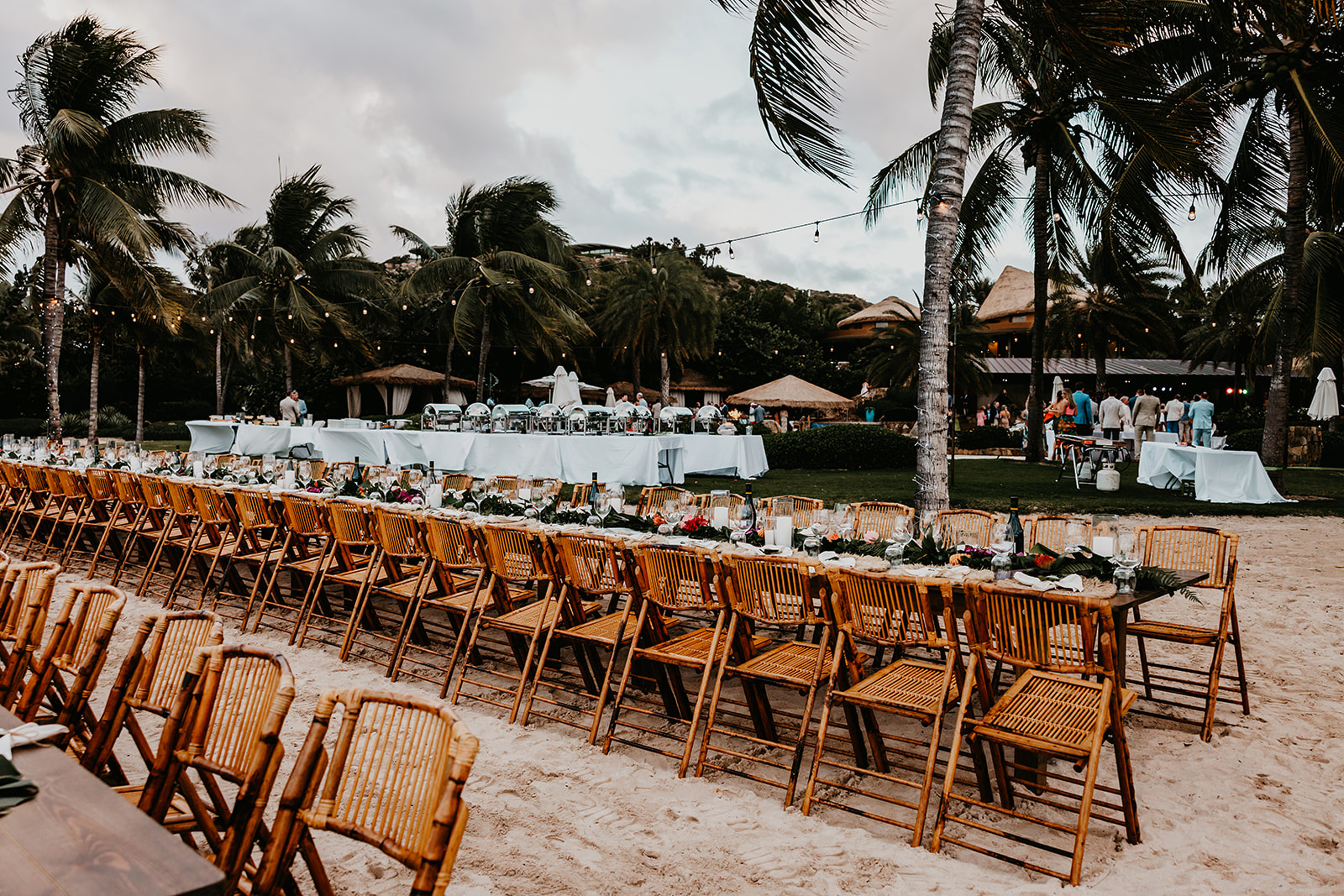 A beach wedding reception setup by BVI Weddings & Events features long wooden tables and chairs arranged in rows on the sand. Tables are decorated with flowers, candles, and greenery. Palm trees and string lights are visible in the background, along with some guests.