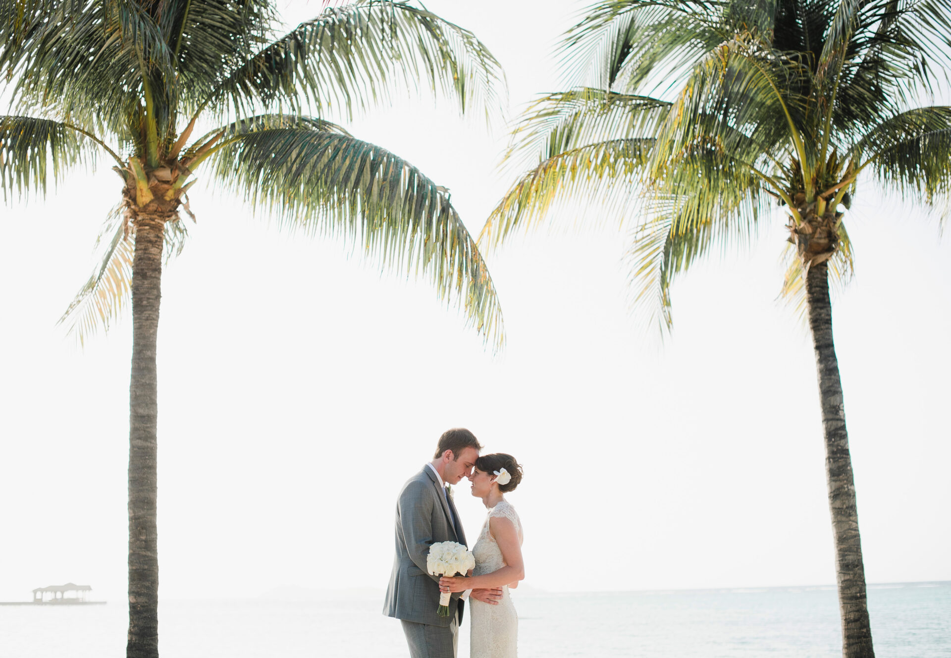 A newlywed couple stands close together, forehead to forehead, between two tall palm trees on a beach. The bride holds a bouquet of white flowers, and the ocean is visible in the background under a bright, clear sky—perfect for BVI Weddings & Events.