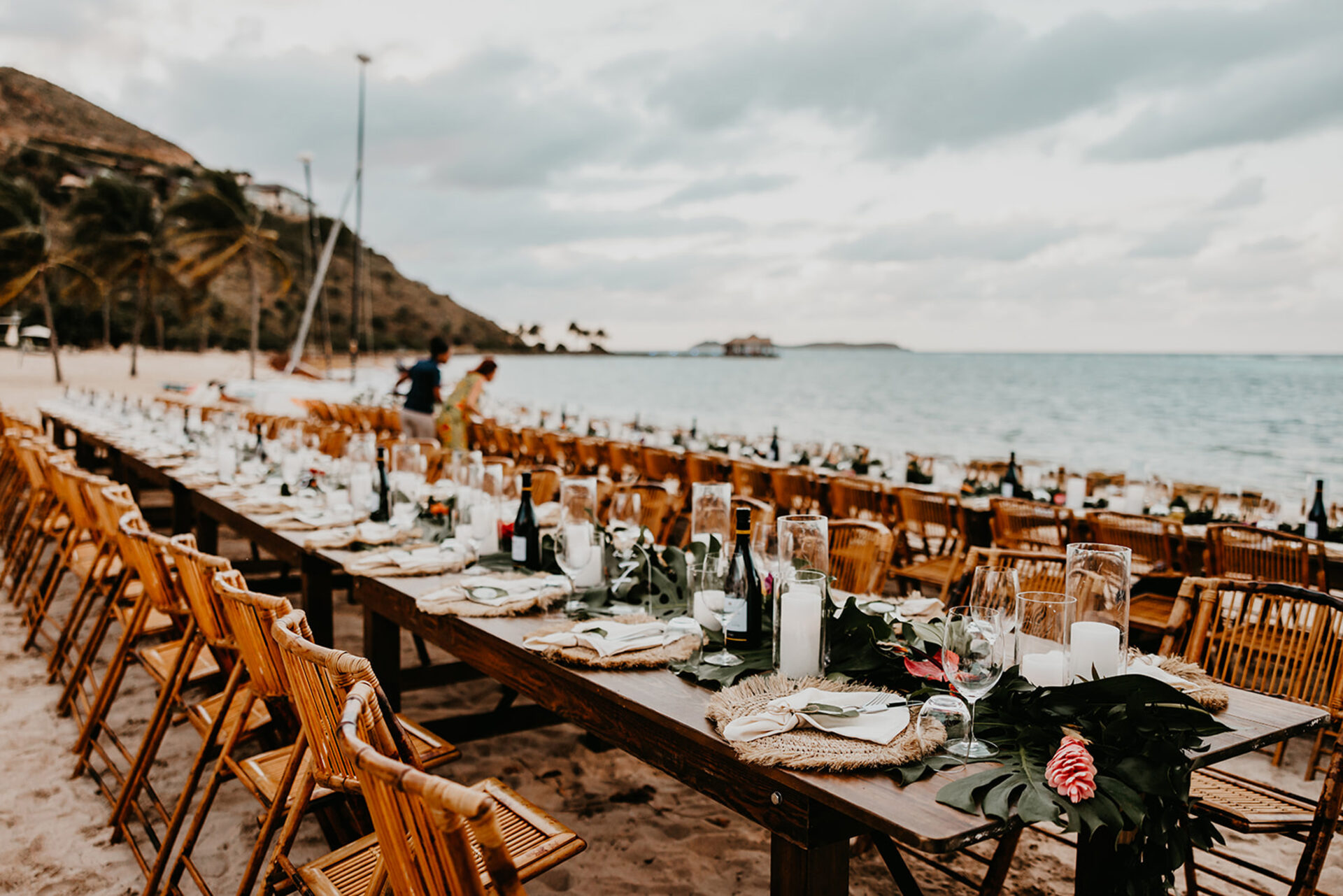 An elegant outdoor dining setup on a beach, curated by BVI Weddings & Events, features long wooden tables adorned with candles, greenery, and floral arrangements. Rows of wooden chairs line the tables, set against a backdrop of palm trees, a cloudy sky, and the ocean.