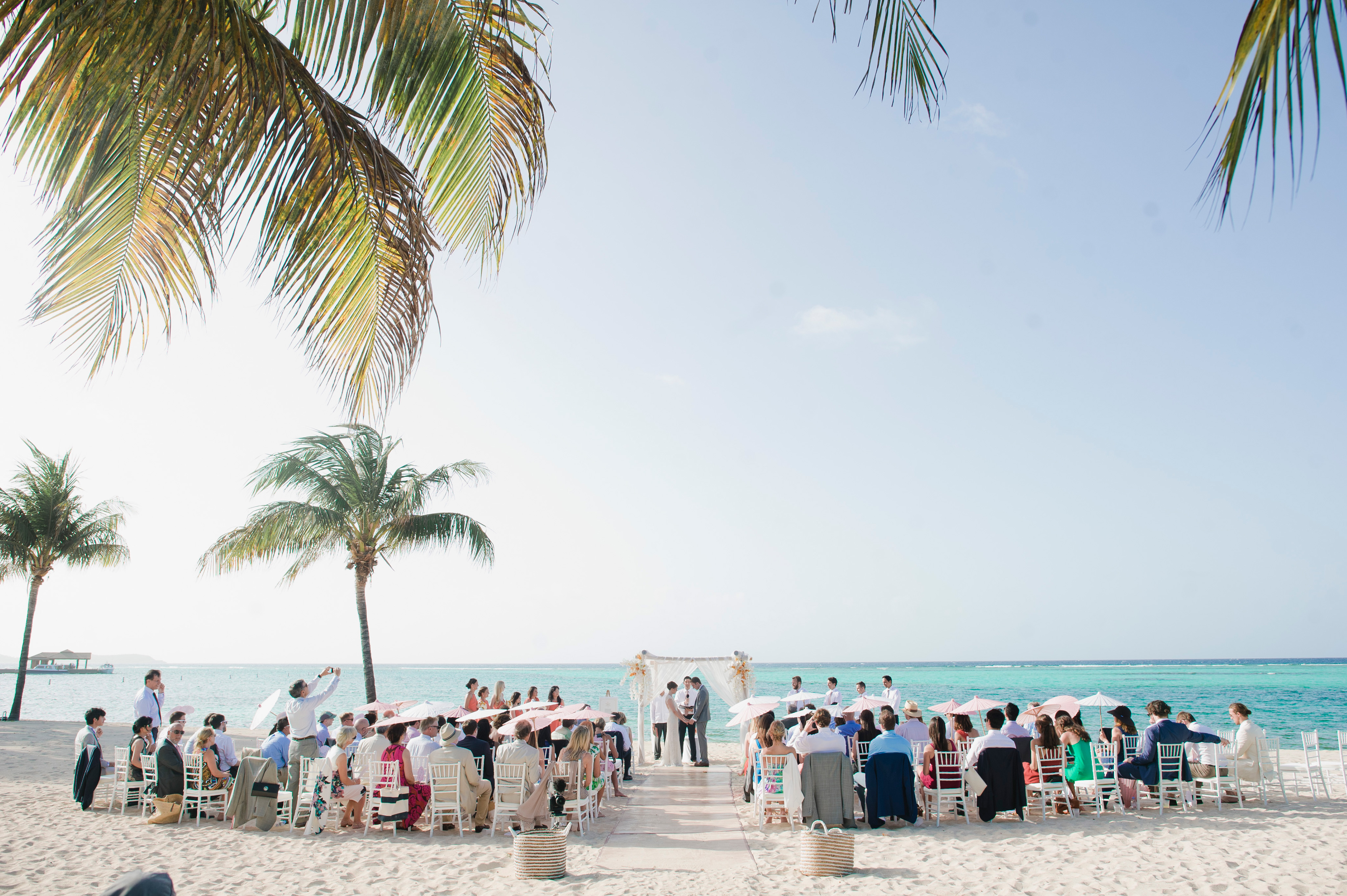 A beach wedding ceremony by BVI Weddings & Events features guests seated on either side of a sandy aisle adorned with white rose petals. The backdrop includes turquoise ocean water, a clear sky, and palm trees. The couple stands under a simple wedding arch decorated with white fabric and flowers.