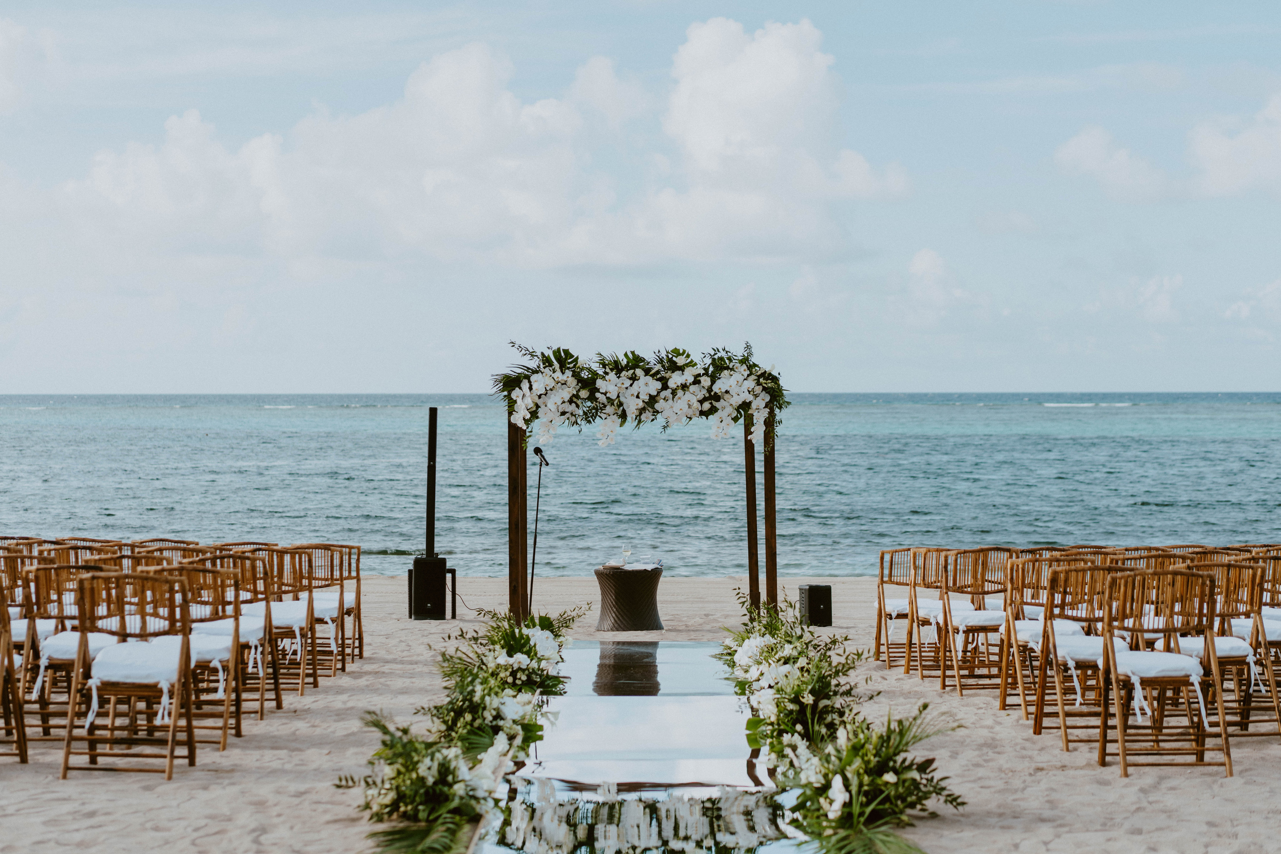 A beautifully decorated beach wedding setup on a sunny day, organized by BVI Weddings & Events. A floral arch stands at the end of an aisle lined with greenery and white flowers, leading to rows of wooden chairs arranged on the sandy shore. The calm ocean and blue sky are in the background.