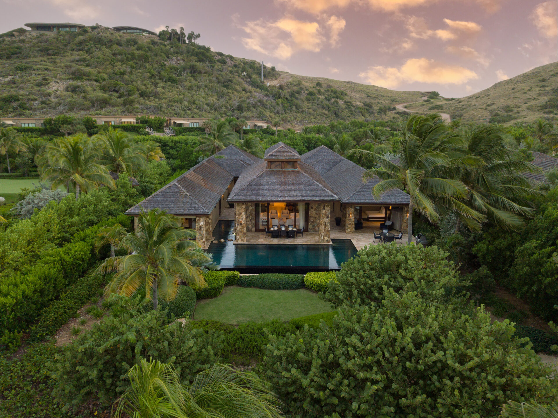 Aerial view of a luxurious Caribbean villa surrounded by lush greenery and trees. The villa has multiple peaked roofs and large windows, and features an infinity pool in the foreground. Rolling hills and a pinkish-orange sunset sky form the scenic background.