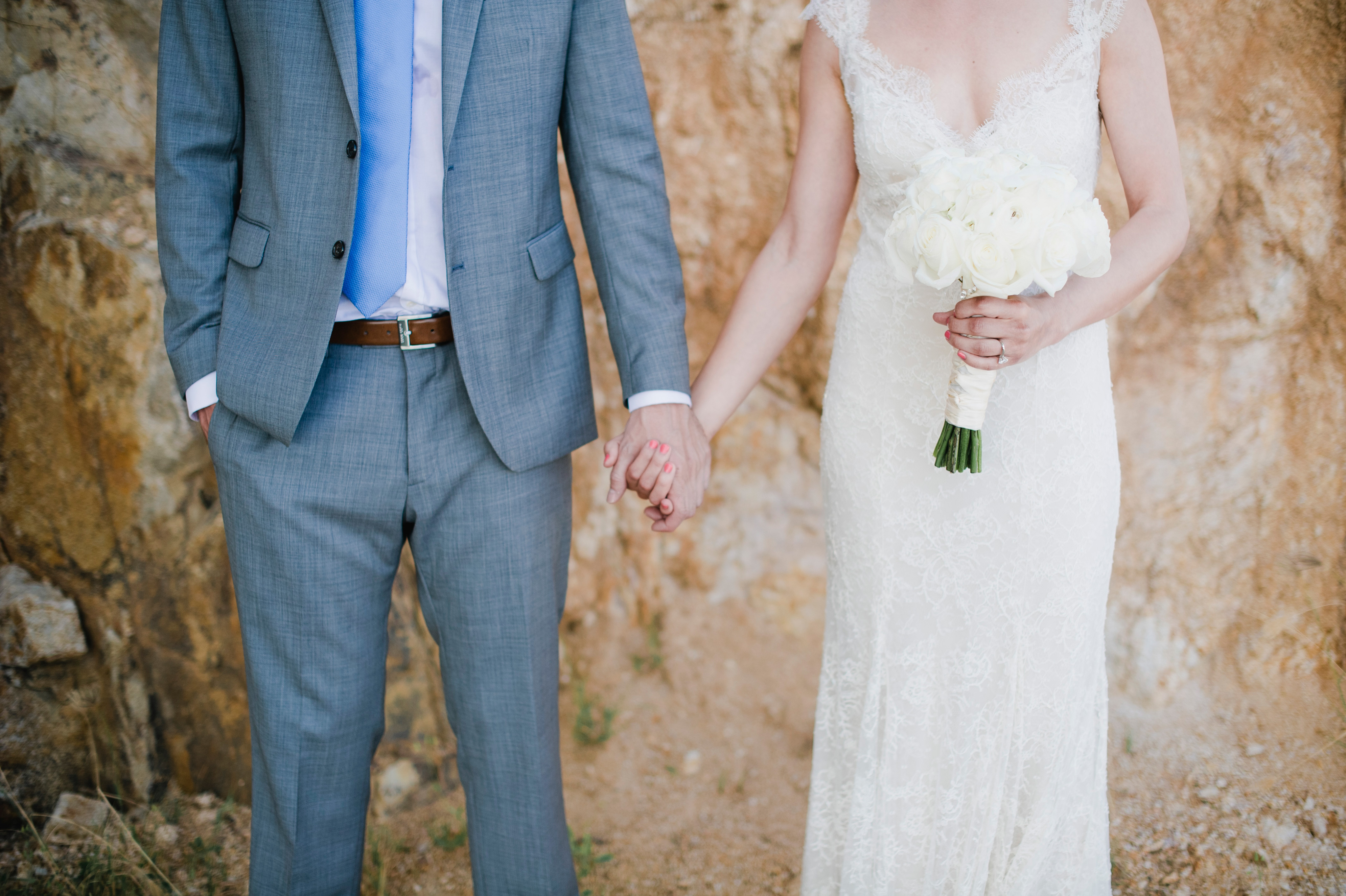 A couple is holding hands. The groom, in a grey suit with a blue tie, and the bride, in a white lace dress holding a bouquet of white flowers, stand before a rocky background. Their heads are not visible in the image. A beautiful moment captured perfectly by BVI Weddings & Events.