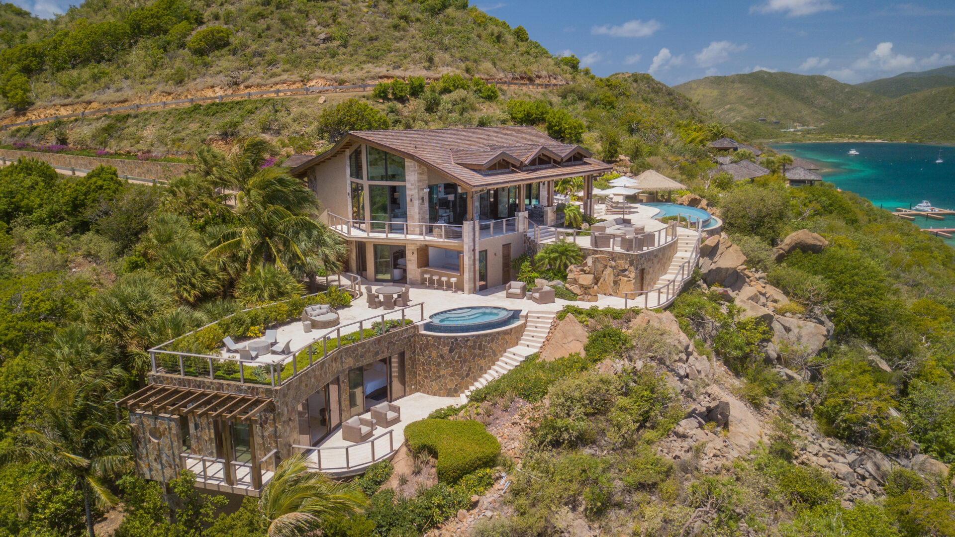 A luxurious, family-friendly villa with multiple terraces and an infinity pool overlooks a lush, green hillside and a turquoise Caribbean bay. The villa includes large windows, outdoor seating areas, palm trees, and a scenic landscape of mountains and water.