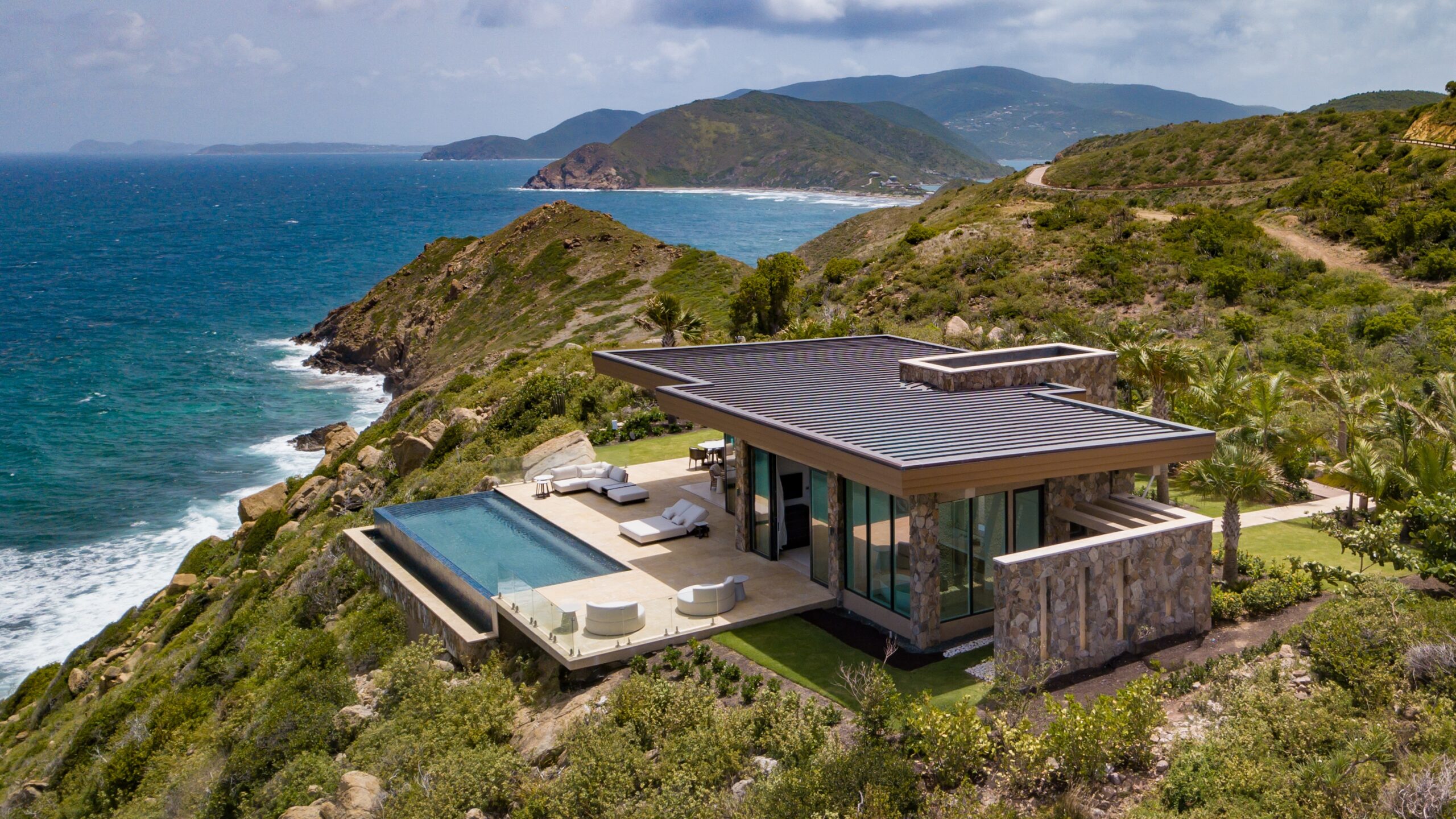 Starlight Villa, a modern house with floor-to-ceiling windows and a flat roof, is perched on a lush green cliff in the Boulder Neighborhood, overlooking a blue ocean. The property features a spacious patio with white outdoor furniture and an infinity pool, with rolling hills and coastal views in the background.