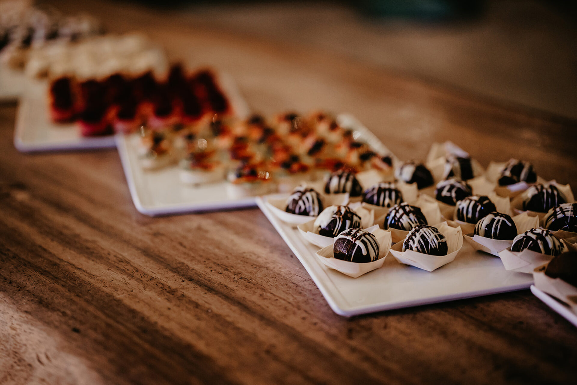 A close-up of several platters of assorted desserts placed on a wooden table. The focus is on chocolate truffles drizzled with white chocolate in the foreground, perfect for BVI Weddings & Events, with an array of other colorful desserts blurred in the background.