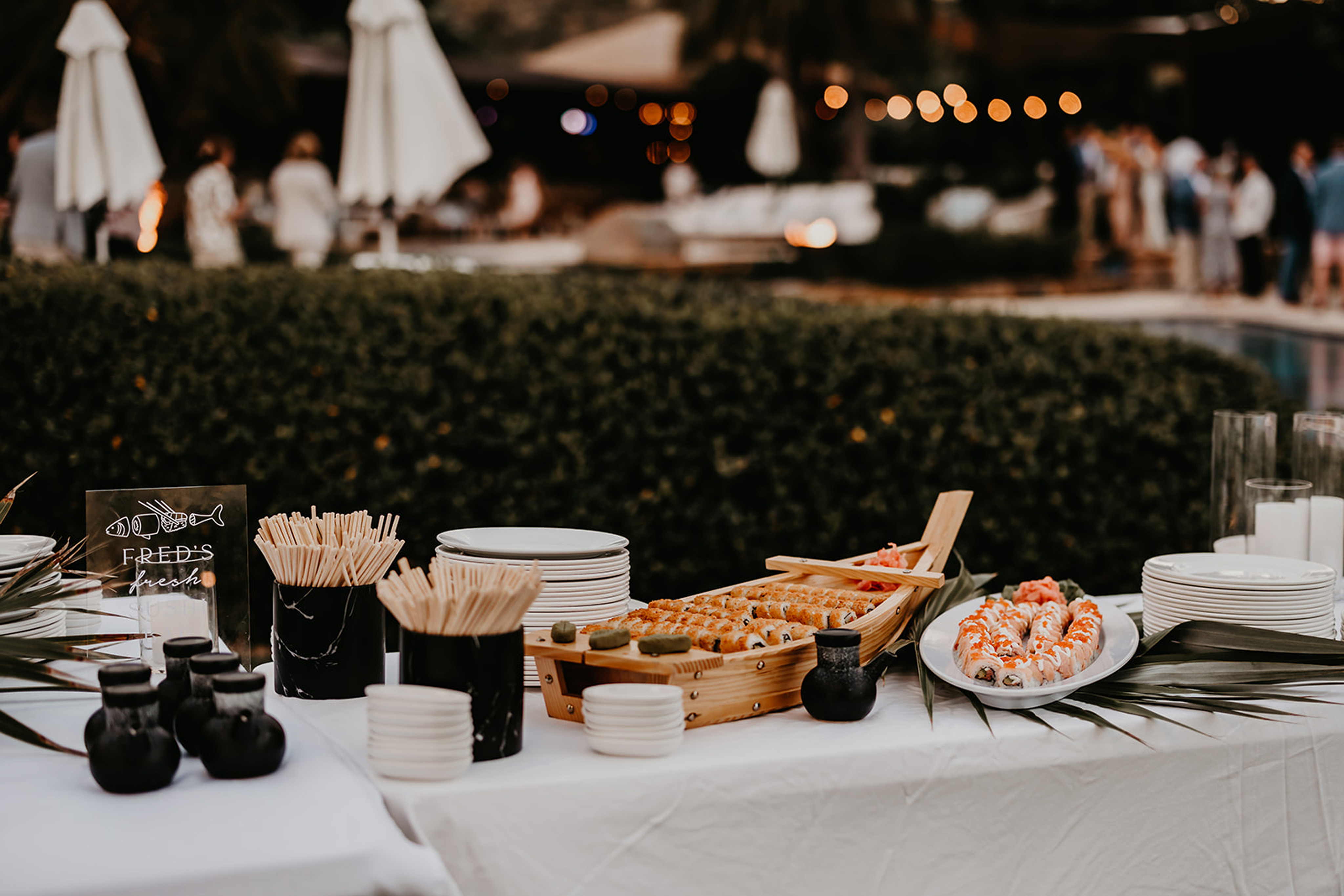 A table set up for an outdoor sushi event by BVI Weddings & Events, featuring plates of sushi rolls, chopsticks, soy sauce bottles, and dipping trays. In the background, people are mingling near white patio umbrellas and greenery.