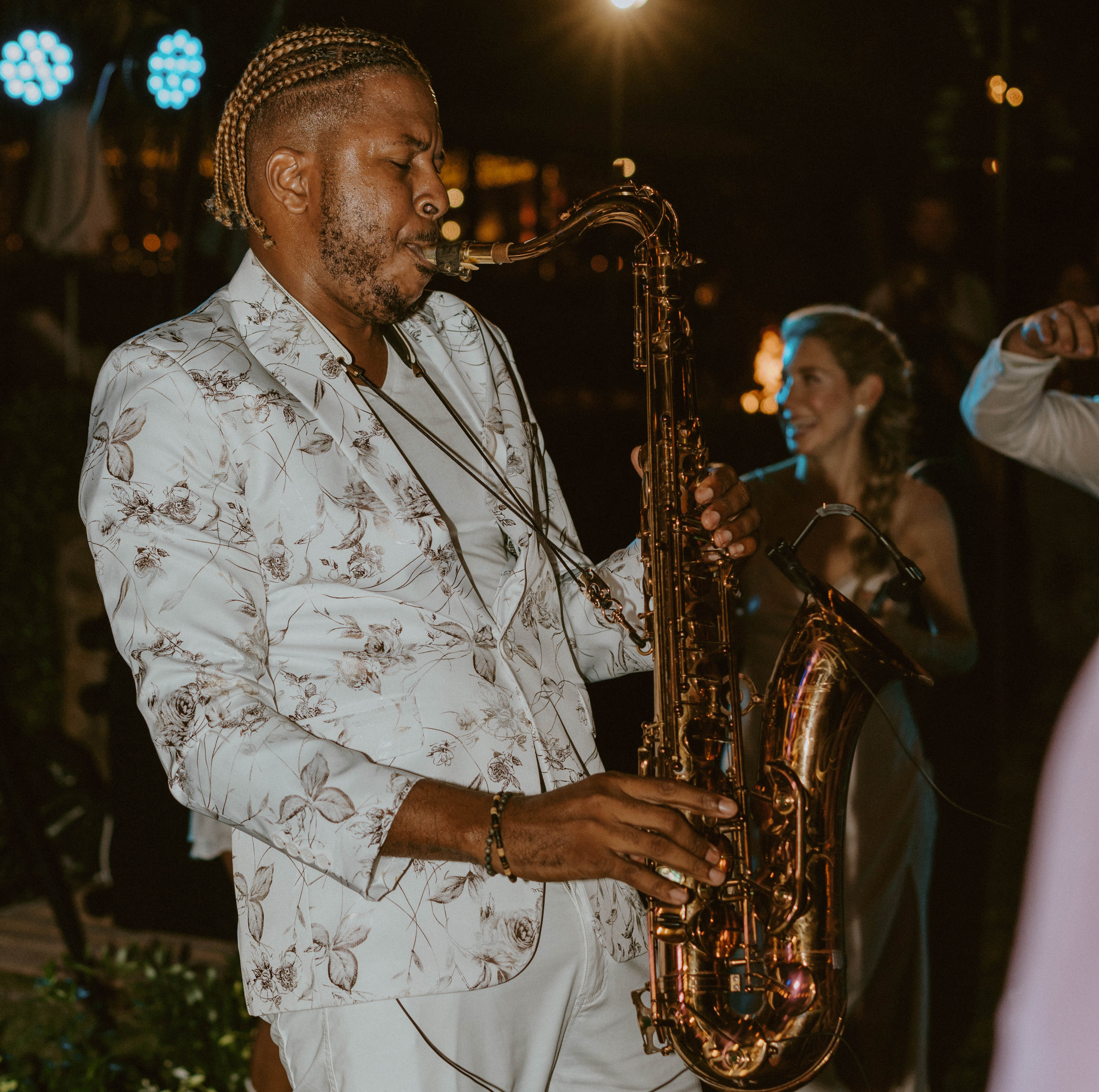 A musician wearing a light-colored floral suit plays the saxophone passionately at an outdoor event. He has braided hair and is focused on his performance. A smiling woman in a dress appears in the background, enjoying the music at a BVI Weddings & Events celebration.