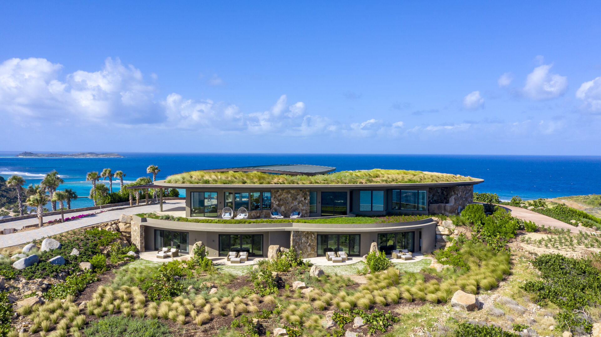 A modern, eco-friendly home with a green roof is situated on a grassy hillside overlooking the vibrant blue ocean in Oil Nut Bay. The house features large windows and outdoor seating areas. Palm trees and lush vegetation surround the property under a clear blue sky with scattered clouds.