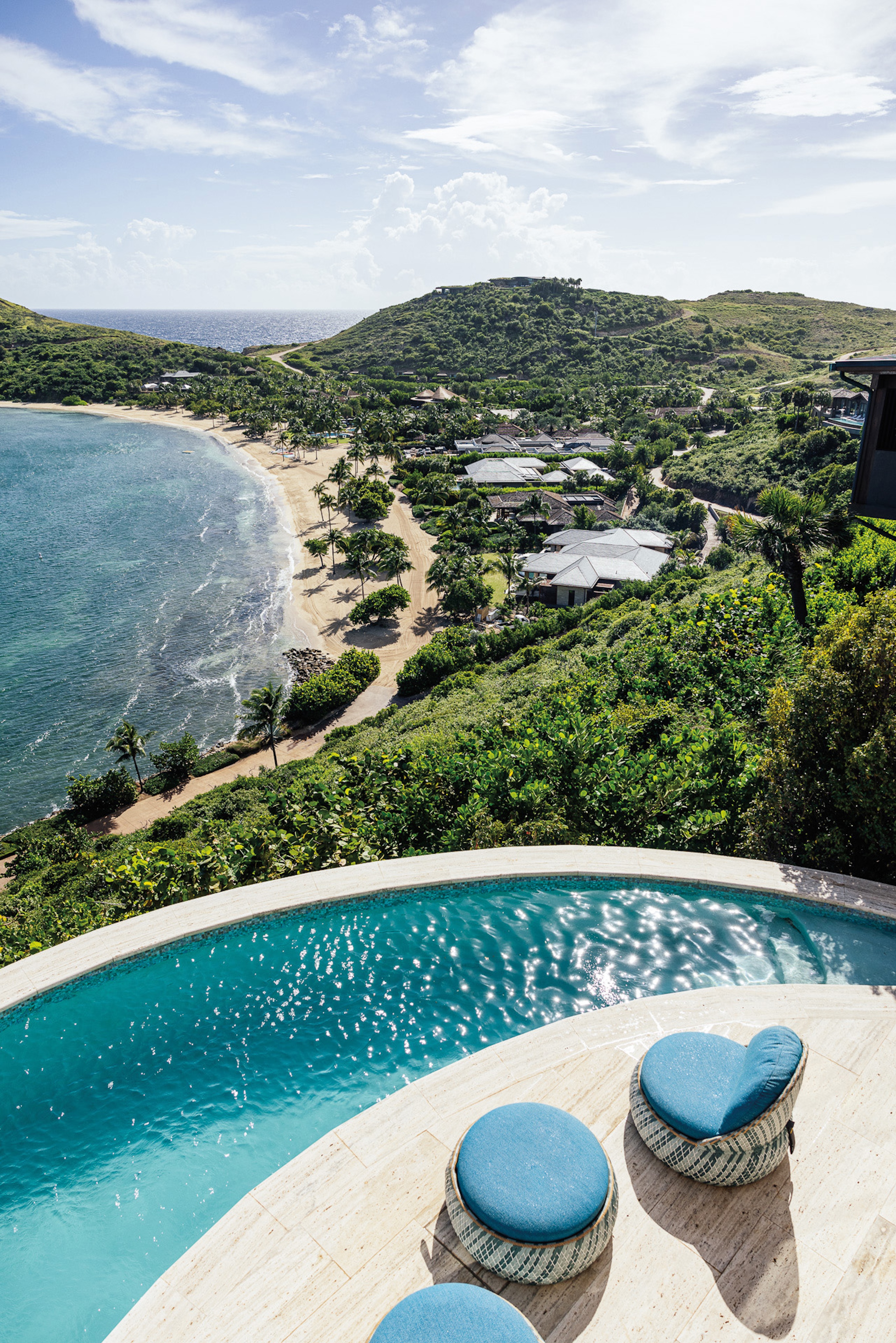 An elevated view overlooking a luxurious round pool with blue lounge chairs, set above a tropical beach. The clear blue ocean meets the sandy shore, surrounded by lush greenery and scattered buildings in the Ridge Neighborhood. Hills covered in vegetation rise in the background under a mostly clear sky.