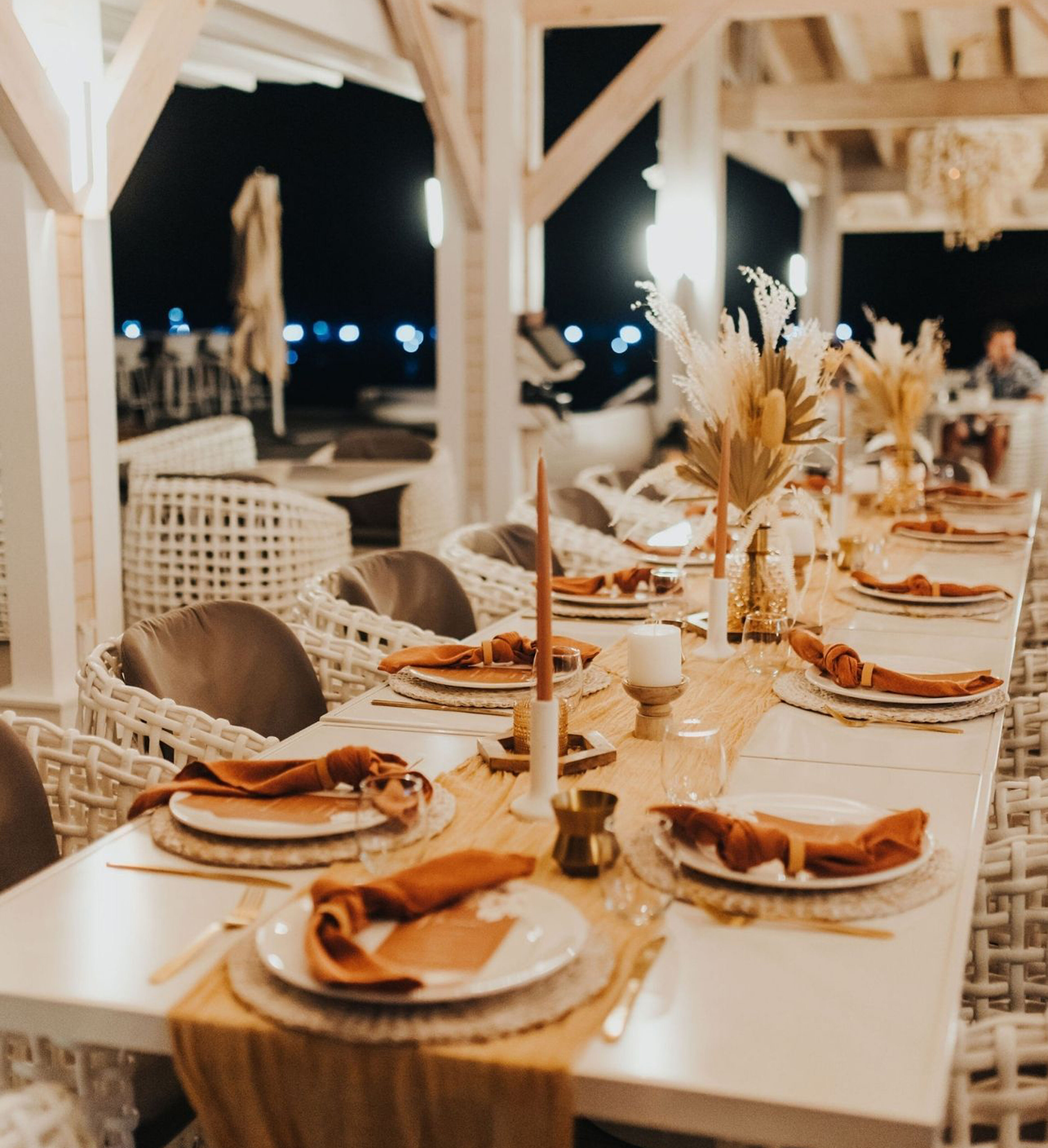 A beautifully decorated dining table set for a dinner party, adorned with plates, silverware, amber-colored napkins, and tall candles. The table runner and floral centerpieces add elegance. Wicker chairs surround the table under a wooden pergola with night lights in the background by BVI Weddings & Events.