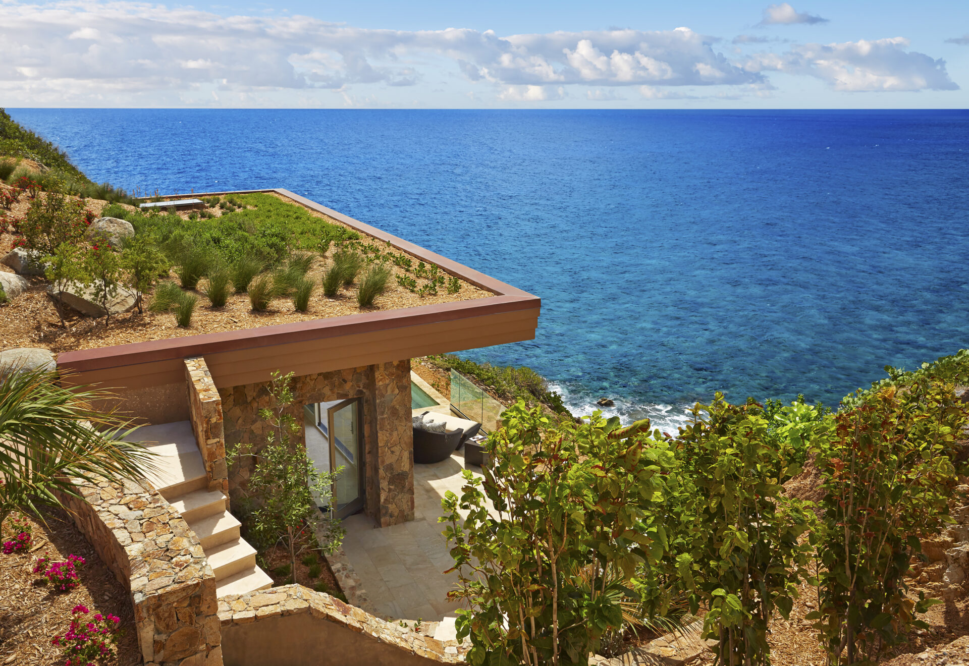 A modern stone house with a rooftop garden is perched on a cliff overlooking the vast, calm ocean of the British Virgin Islands under a partly cloudy sky. Steps and greenery surround the house, blending it into the natural landscape of this most exclusive community in the Caribbean.