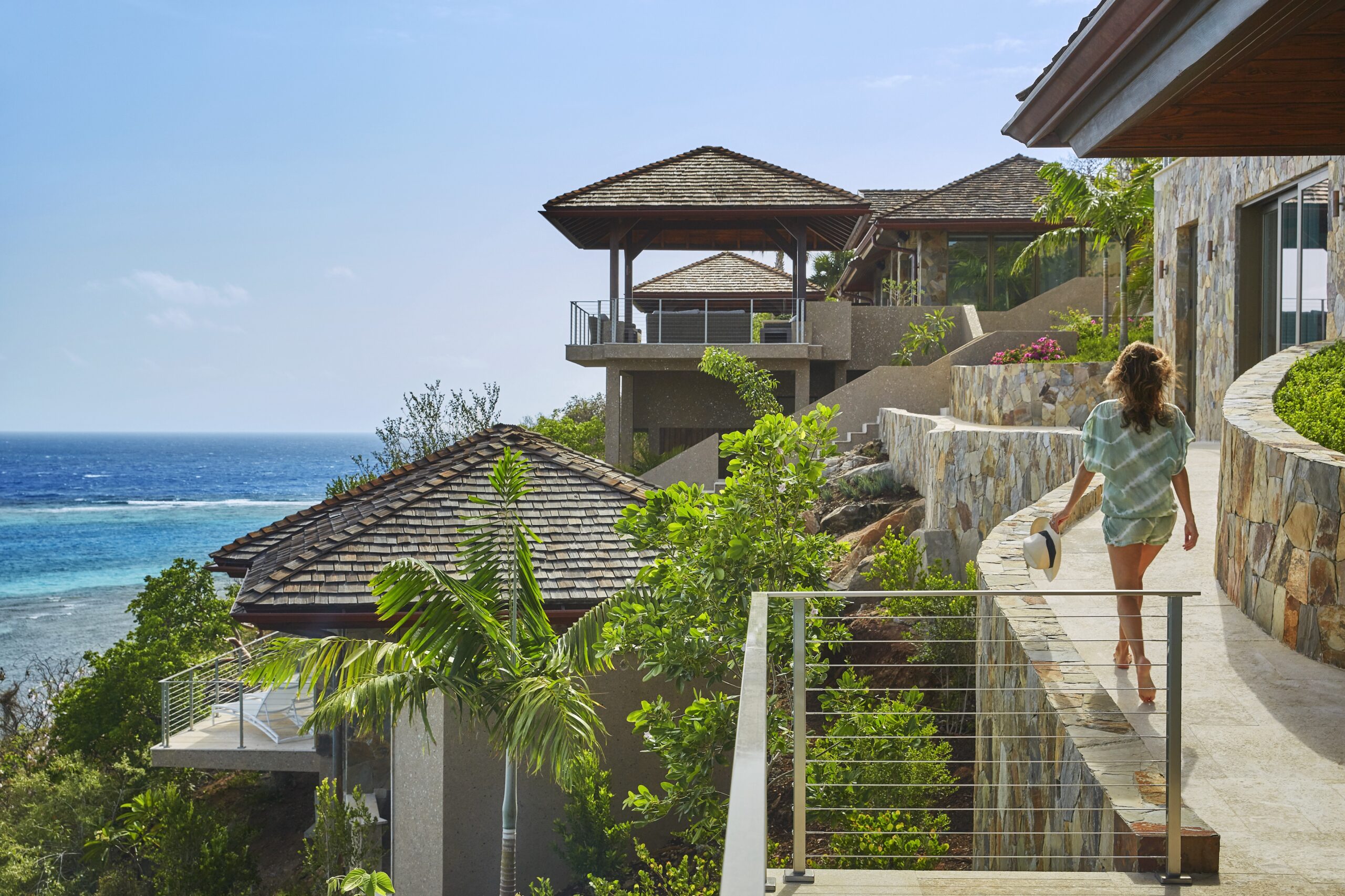 A woman with long hair walks along a stone pathway surrounded by tropical foliage. She carries a hat and towel, heading towards the luxurious villas of Reef House with wooden roofs, overlooking a scenic ocean view under a clear blue sky.