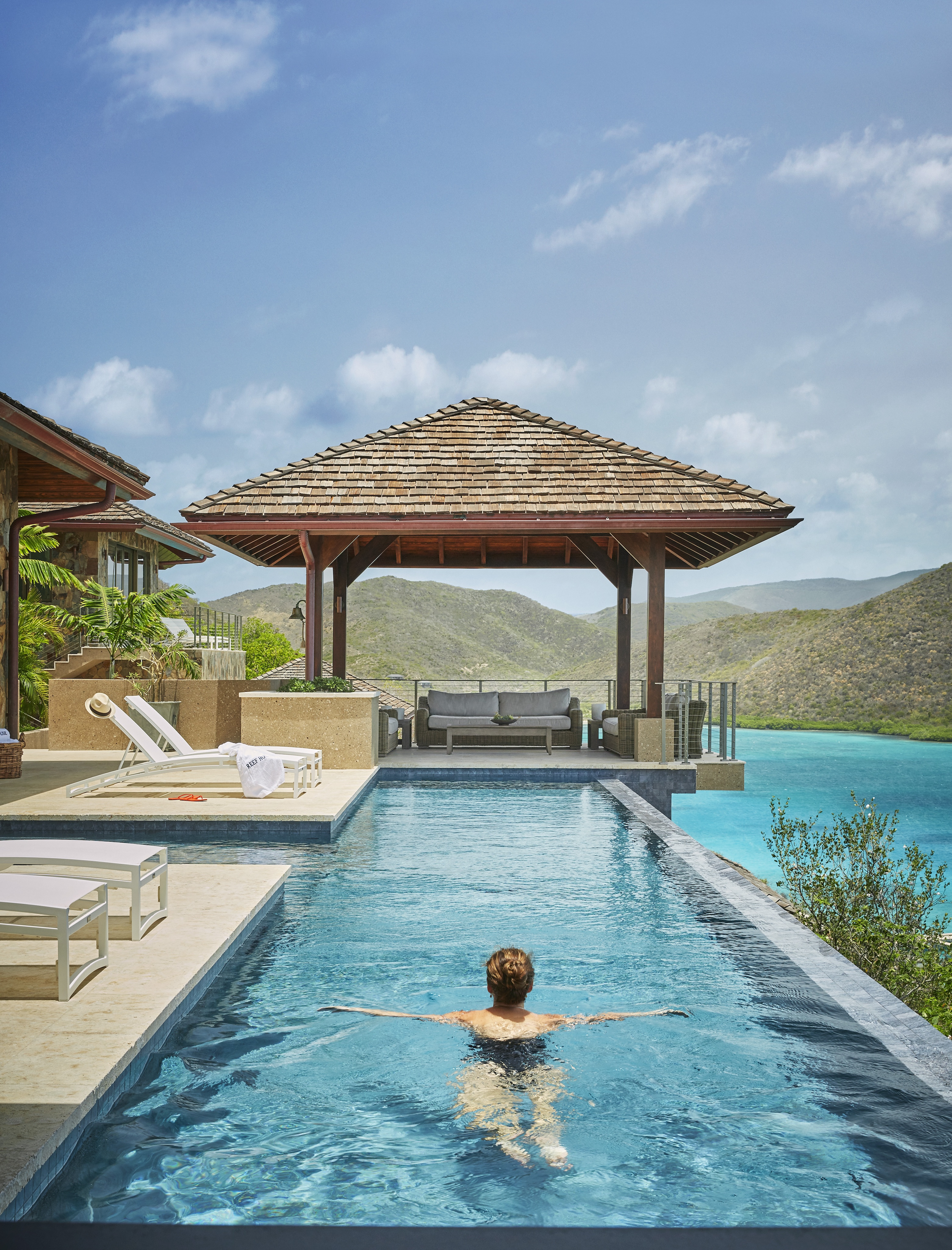 A person swims in an infinity pool overlooking a serene body of water and lush, green hills under a clear blue sky. In the background, there is a shaded pavilion with a small seating area and a lounge chair on the poolside patio, highlighting the elegance of this exclusive Estate Neighborhood.