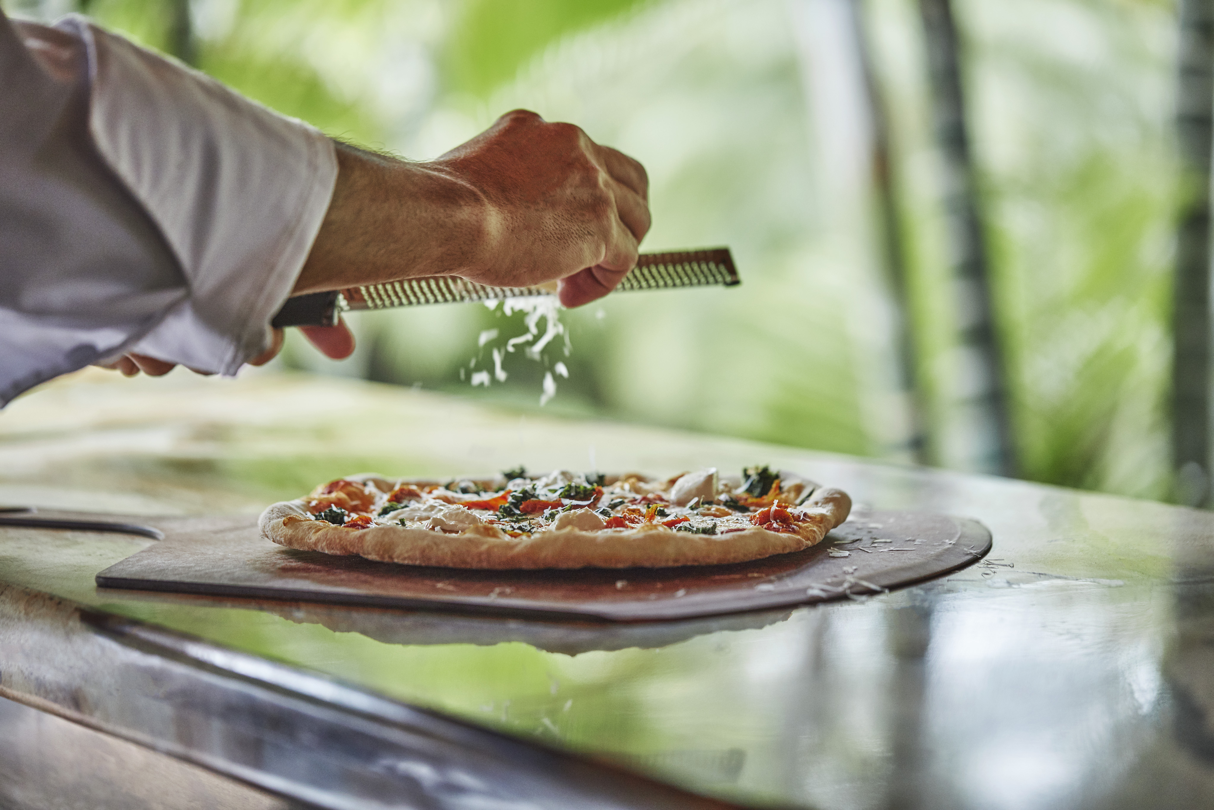 A chef's hands grating cheese over a freshly made pizza on a wooden board. The background is blurred with a lush, green, natural setting reminiscent of Island Dining in the British Virgin Islands. The pizza is topped with various herbs and vegetables.