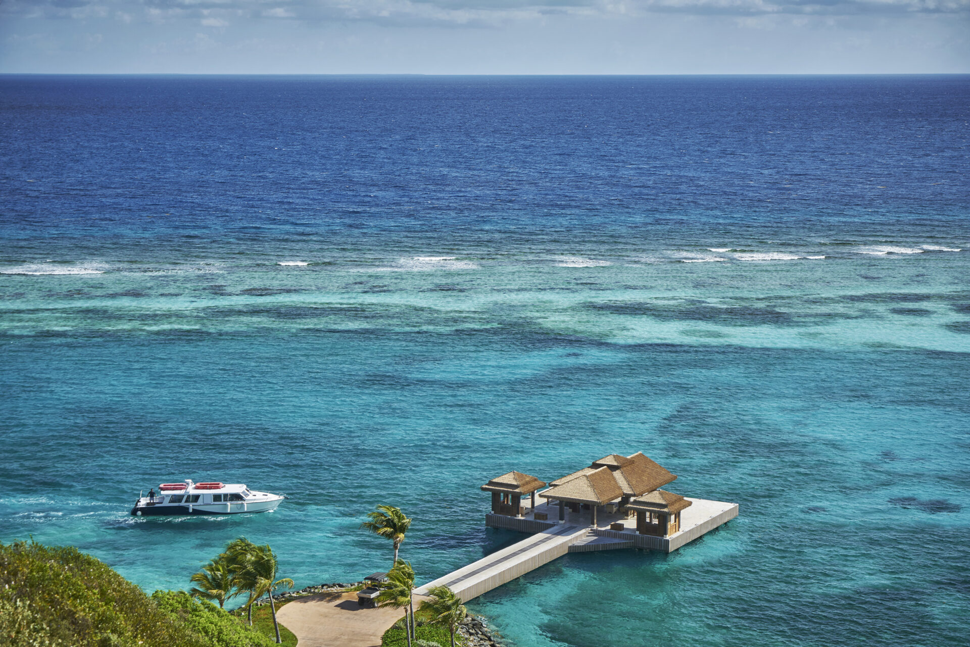 A white boat with red canopies navigates towards a small, wooden dock surrounded by clear, turquoise waters. The dock features a thatched-roof hut. Lush greenery is visible in the foreground, with the open sea and a blue sky in the background—a perfect setting for luxury honeymoons at all-inclusive Caribbean resorts.