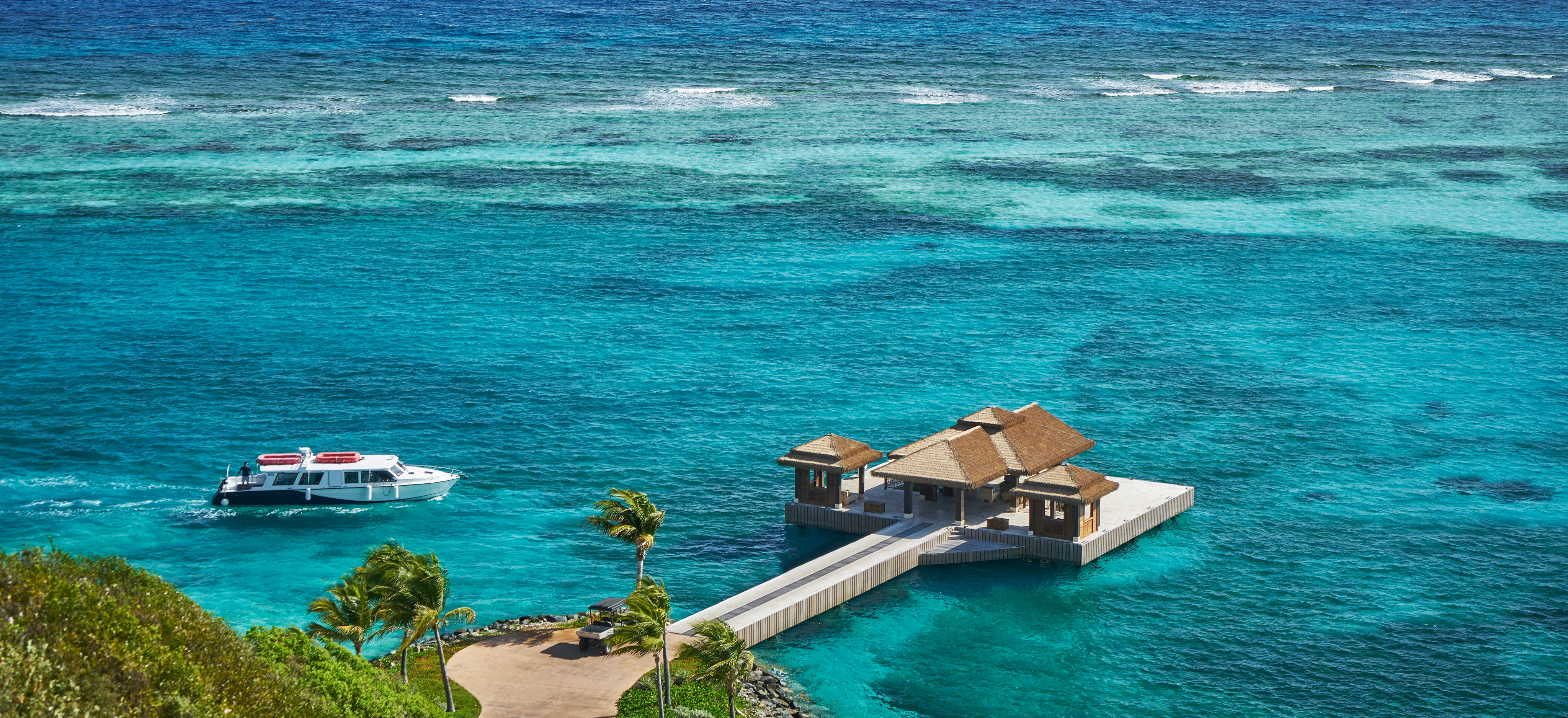 A boat approaches a small, secluded pavilion built on a turquoise blue ocean with a concrete path leading from the pavilion to the lush green coastline. Part of the luxury real estate property for sale in Oil Nut Bay, British Virgin Islands, it offers stunning views and varying shades of blue water with small waves visible in the distance.